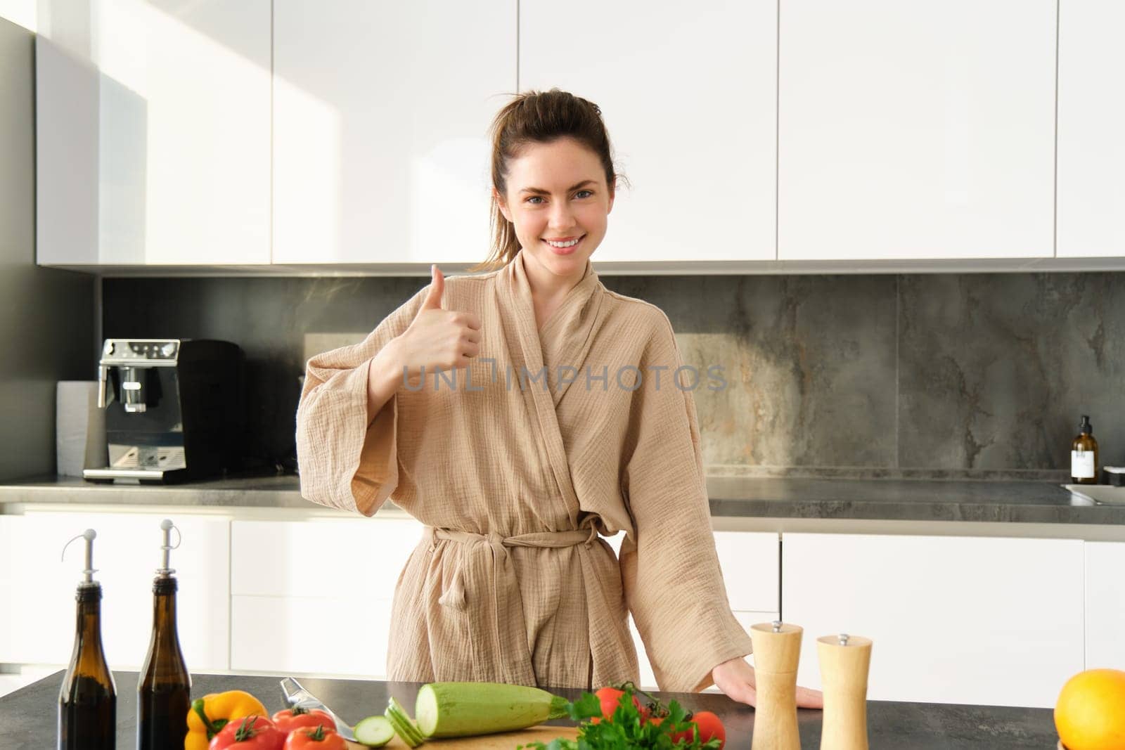 Portrait of happy young woman preparing vegetarian dinner, showing thumbs up, wife cooking in kitchen and smiling, recommending product, chopping vegetables for healthy salad.