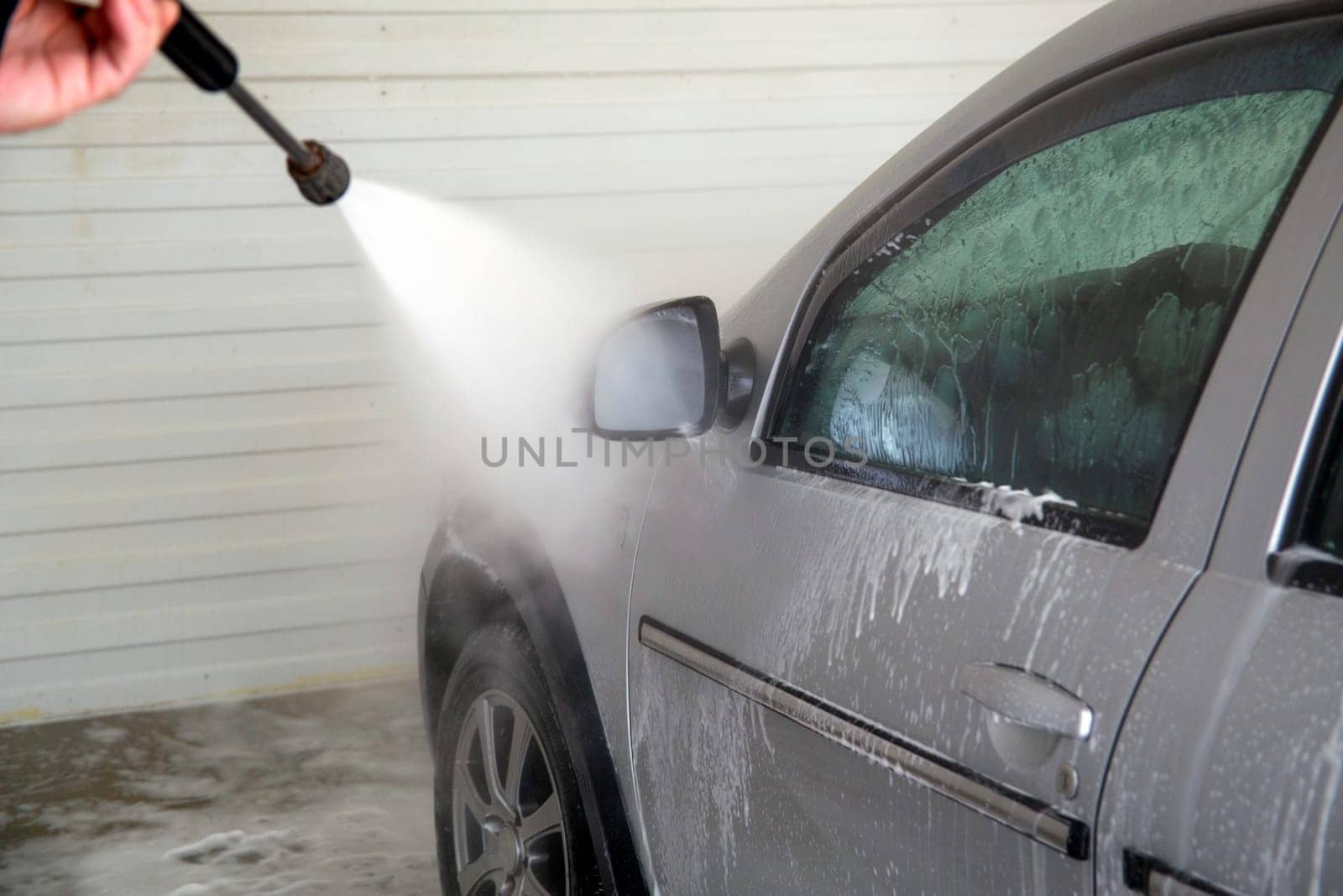 Indoor car washing. Process of removing soap foam from side of vehicle with pressure washer - close-up with selective focus.
