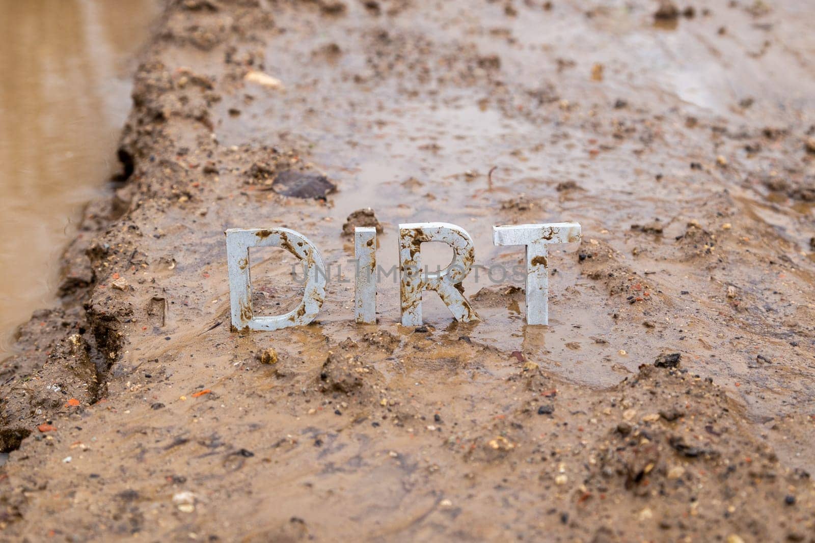 the word dirt composed of silver metal letters on wet clay surface with selective focus, background blur and linear perspective in cloudy autumn day light