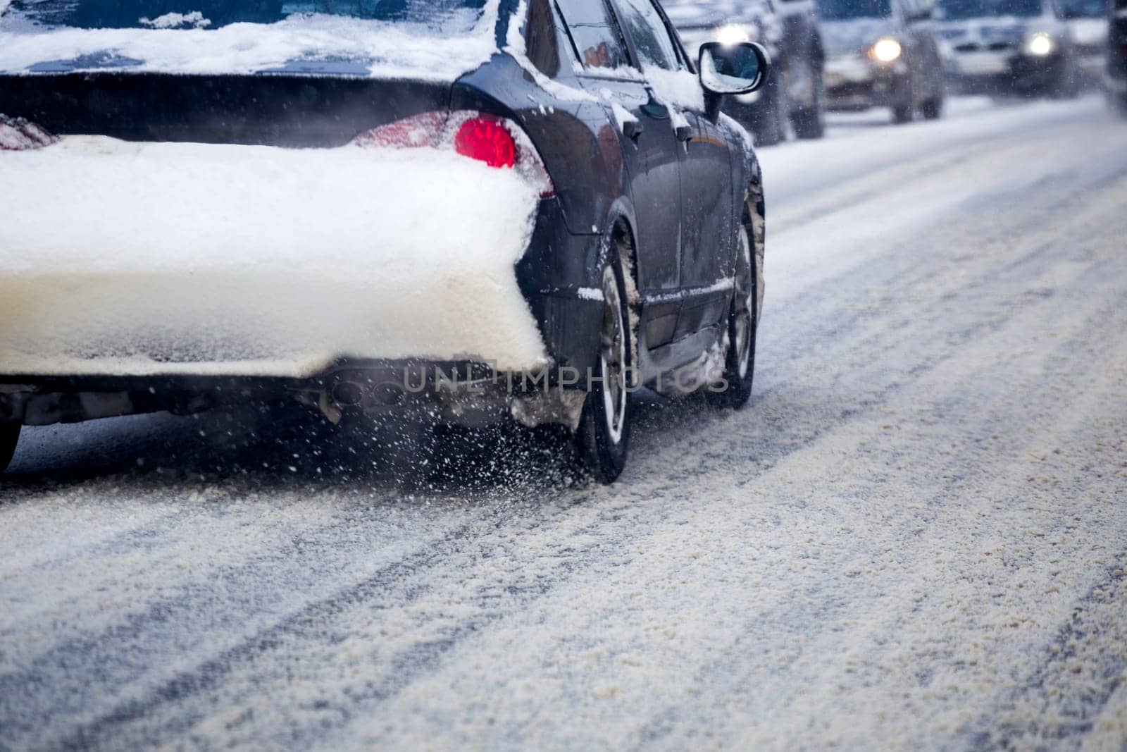 Road snow flinders flow from wheels of black car moving fast in daylight city with selective focus.