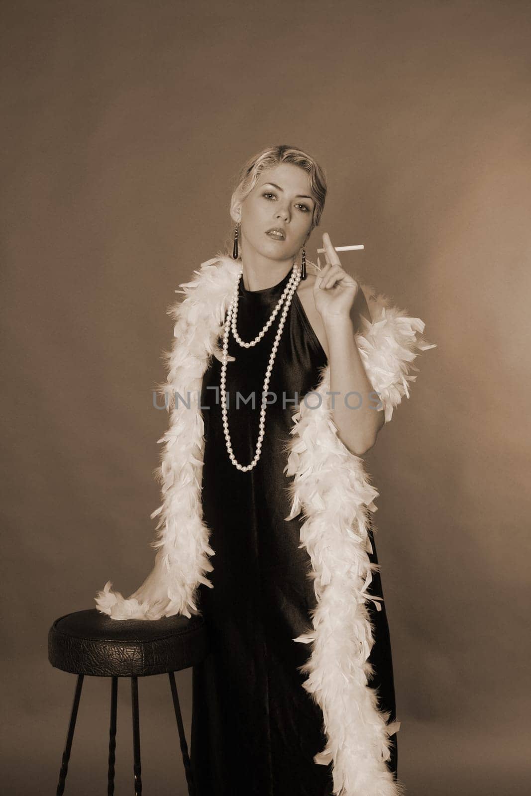 A woman dressed in the style of the 20s stands next to a bar stool with a cigarette.