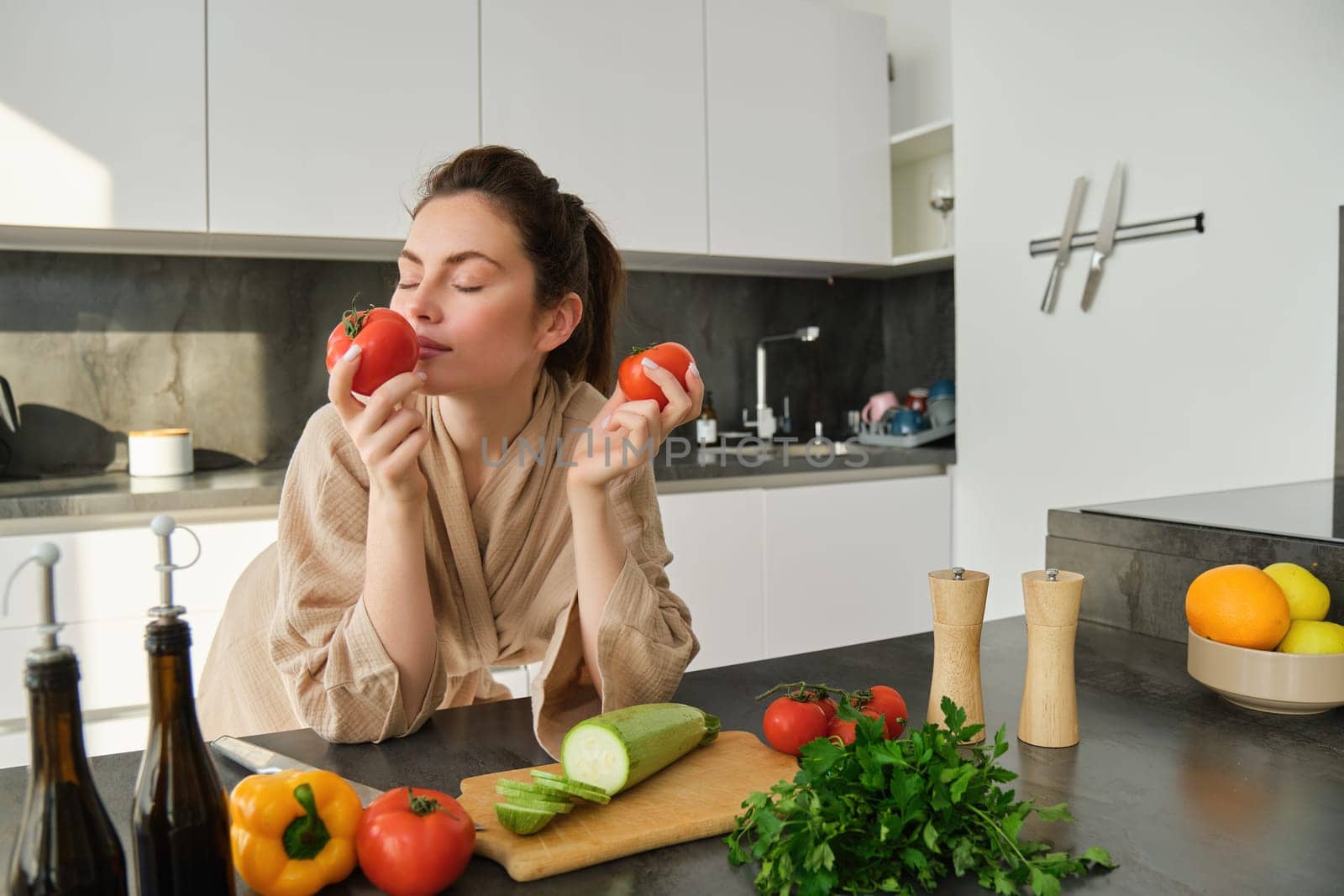 Portrait of beautiful young woman smelling tomatoes, cooking salad from fresh vegetables bought in market, preparing food for family, lead active and healthy lifestyle, standing in kitchen by Benzoix