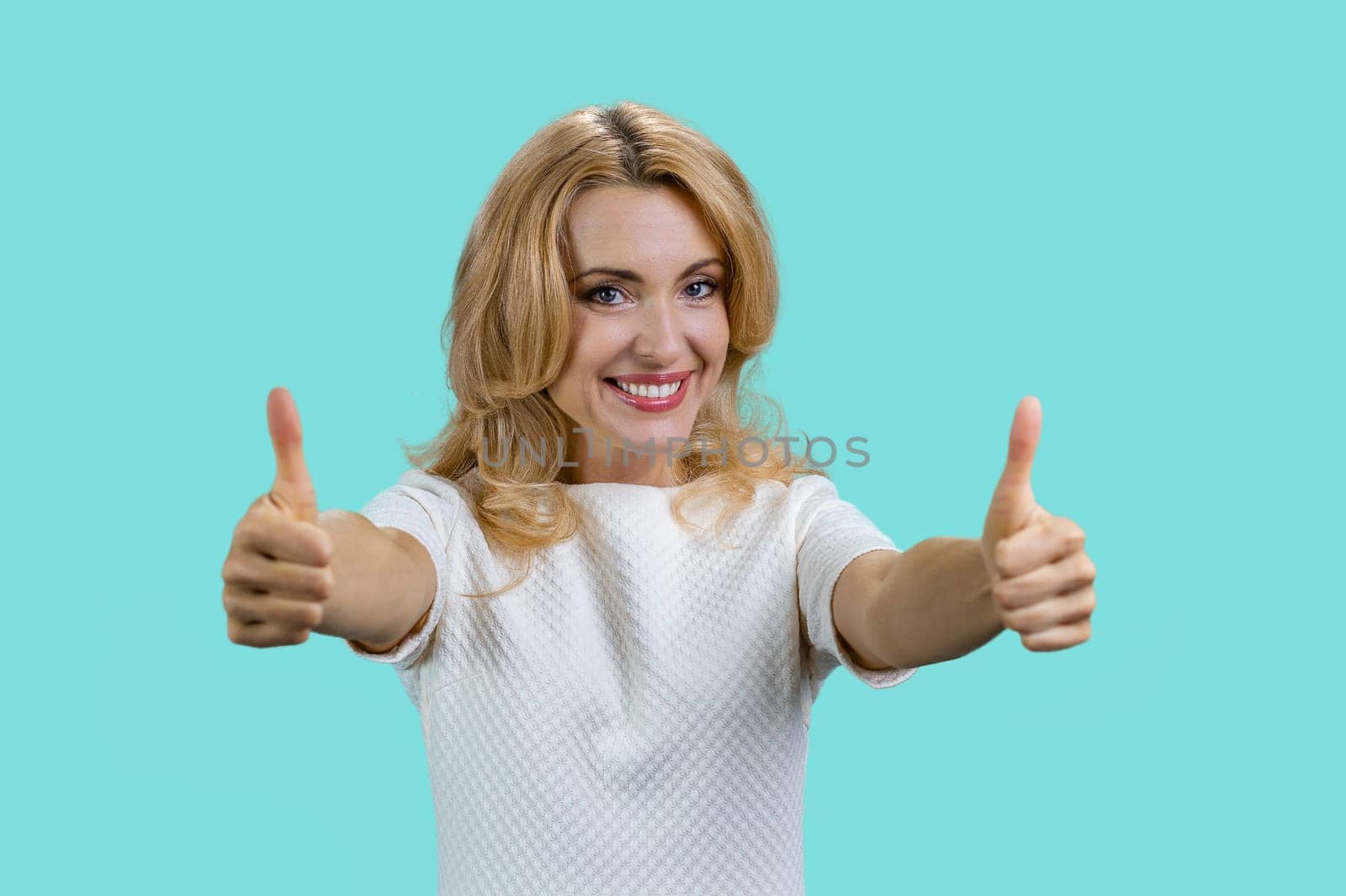 Portrait of a smiling blond woman gives both thumbs up. Isolated on turquoise.