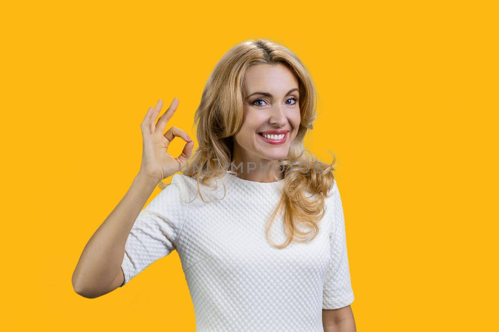 Portrait of a happy mature blond woman shows ok gesture sign. Isolated on orange.