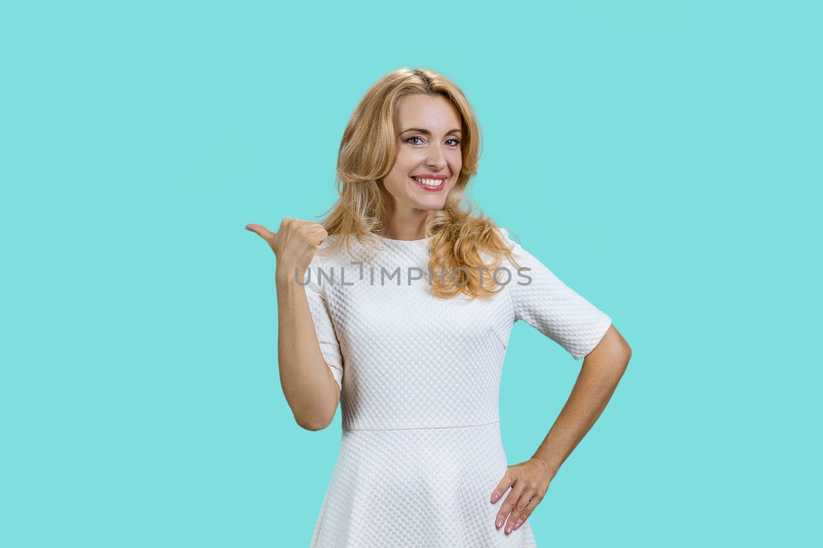 Cheerful mature blond woman pointing back with her thumb. Isolated on turquoise.