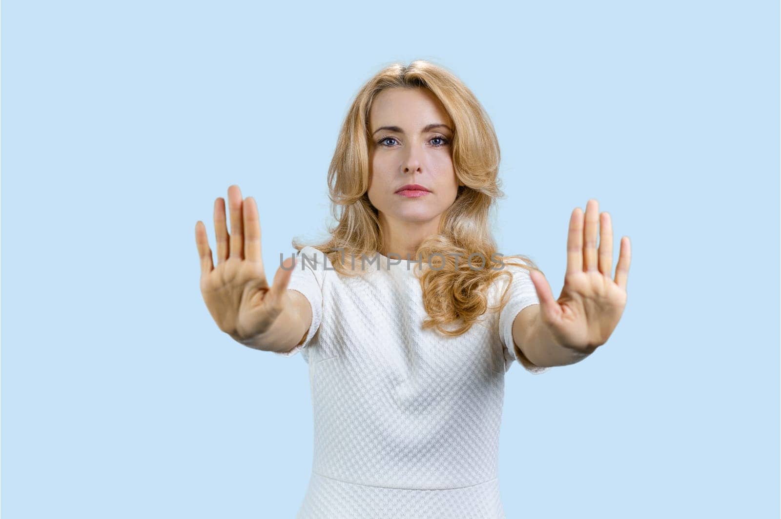 Portrait of caucasian mature woman shows stop sign prohibition with both hands. Isolated on pale blue.