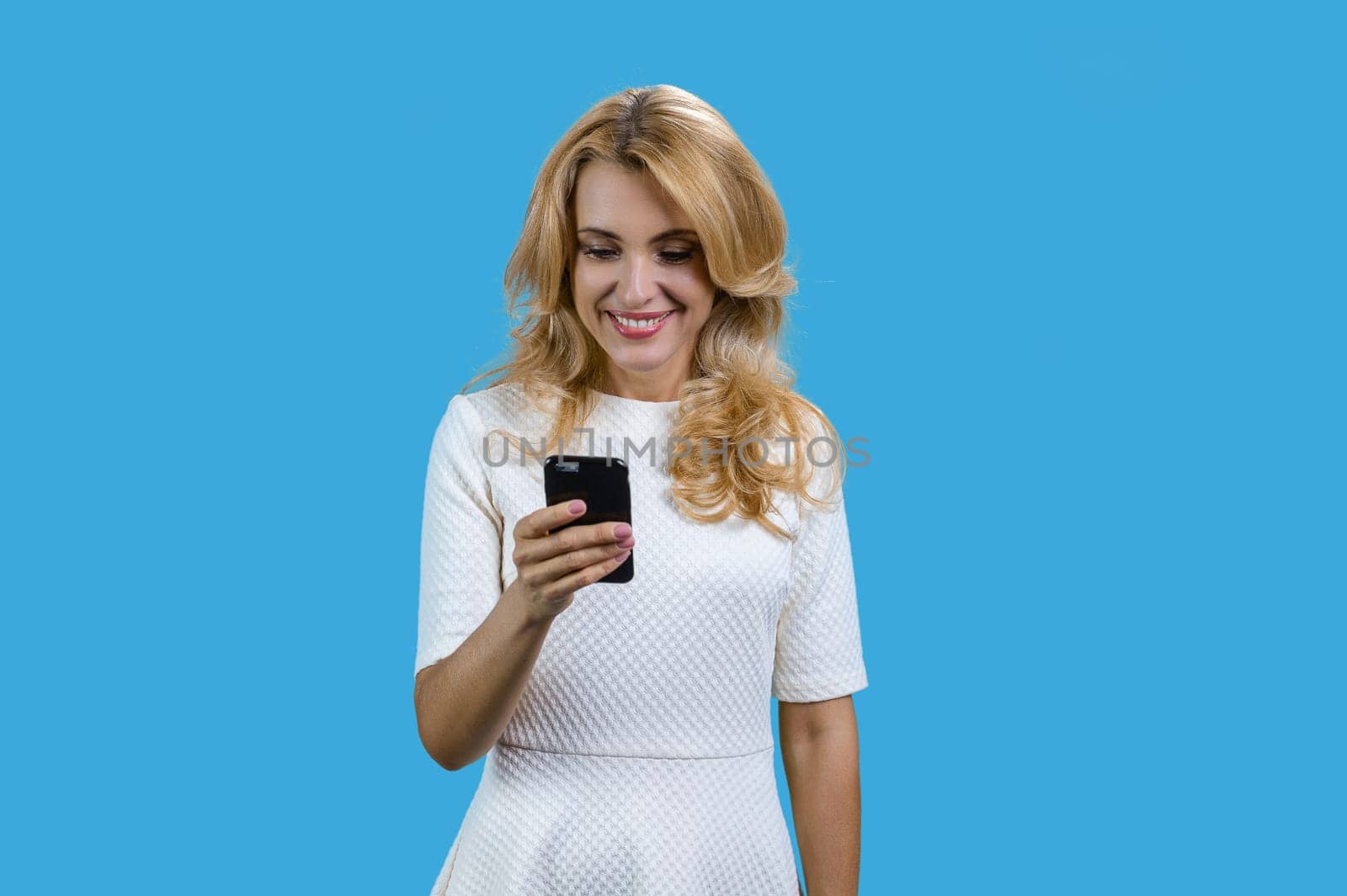 Portrait of happy blonde mature woman using her smartphone. Isolated on blue.