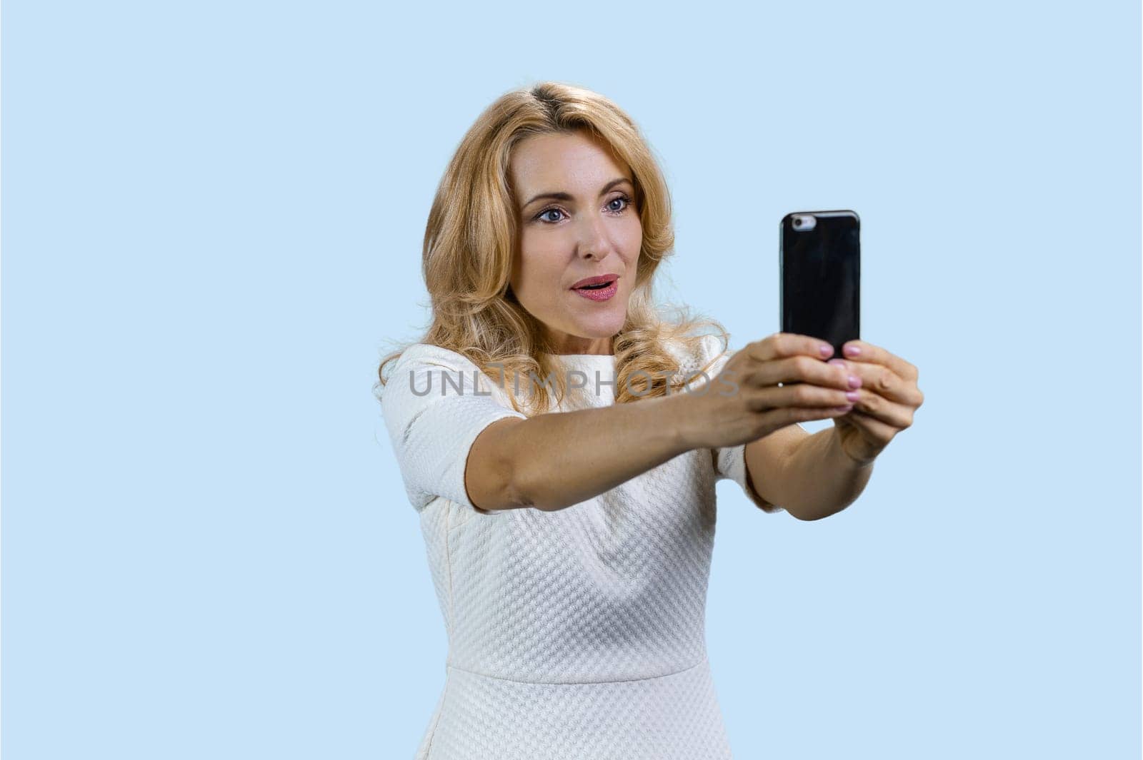 Expressive blonde woman taking selfie on smartphone frontal camera. Isolated pastel blue background.