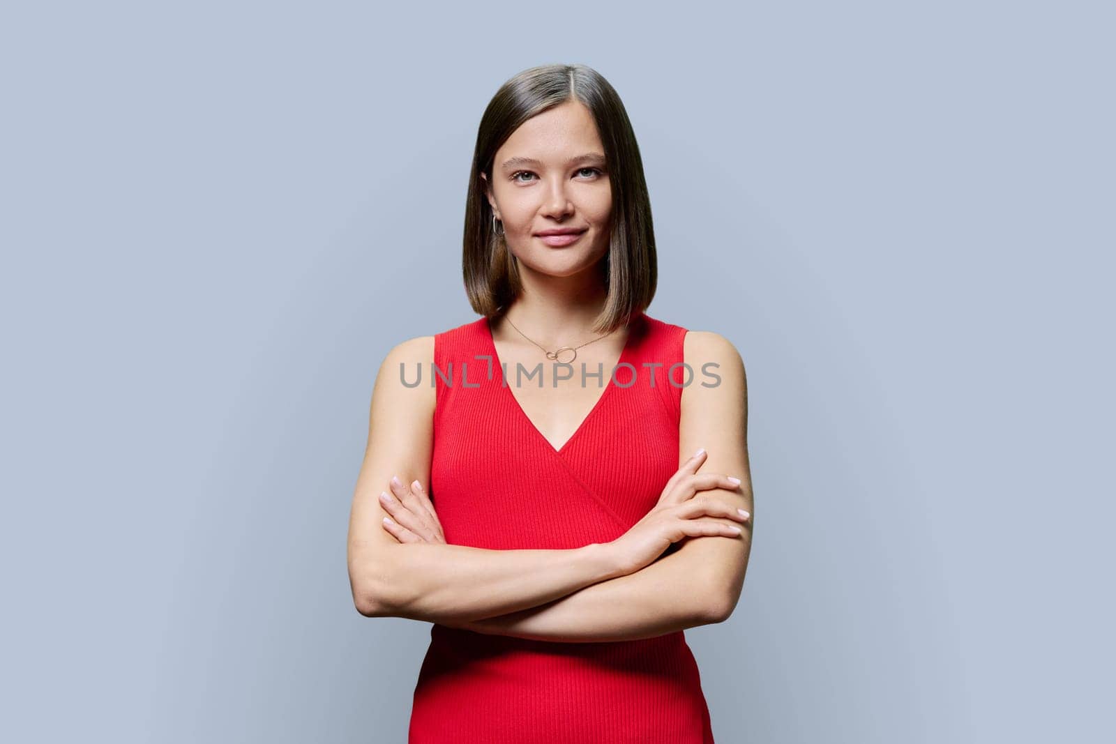 Portrait of young confident woman in red on grey studio background. Successful fashionable female with crossed arms looking at camera. Business, work, services, education, fashion beauty professions