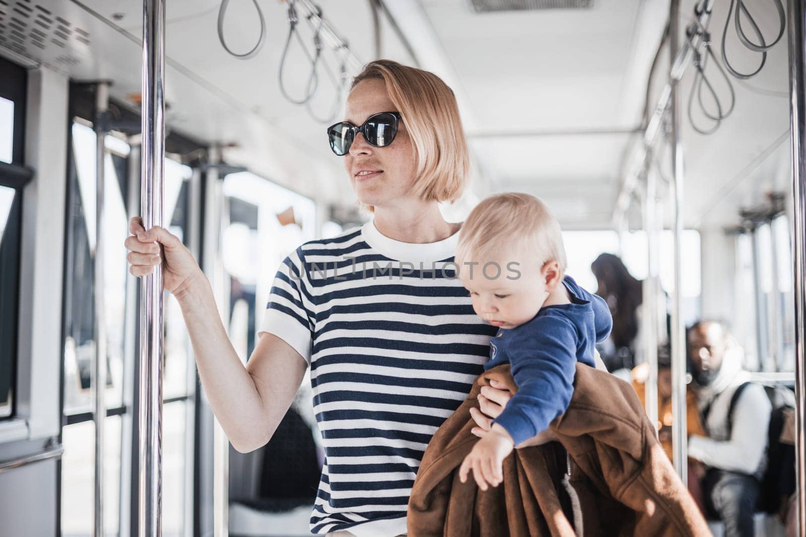 Mother carries her child while standing and holding on to bar holder on bus. Mom holding infant baby boy in her arms while riding in public transportation. Cute toddler traveling with mother