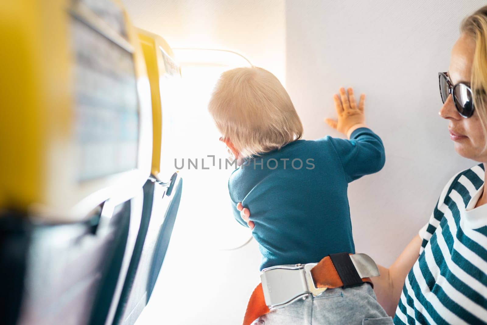 Mom and child flying by plane. Mother holding and playing with her infant baby boy child in her lap during economy comercial flight. Concept photo of air travel with baby. Real people. by kasto