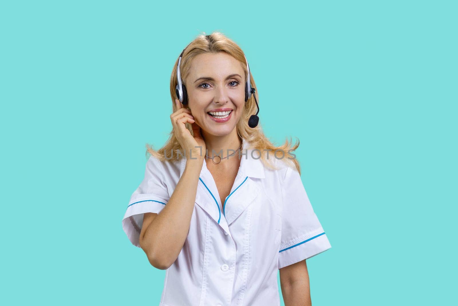 Portrait of happy blonde female doctor wearing headset. Isolated on turquoise.