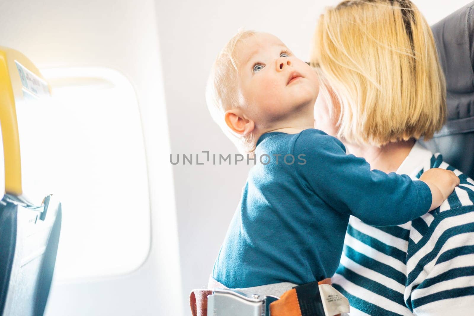 Mom and child flying by plane. Mother holding and playing with her infant baby boy child in her lap during economy comercial flight. Concept photo of air travel with baby. Real people. by kasto