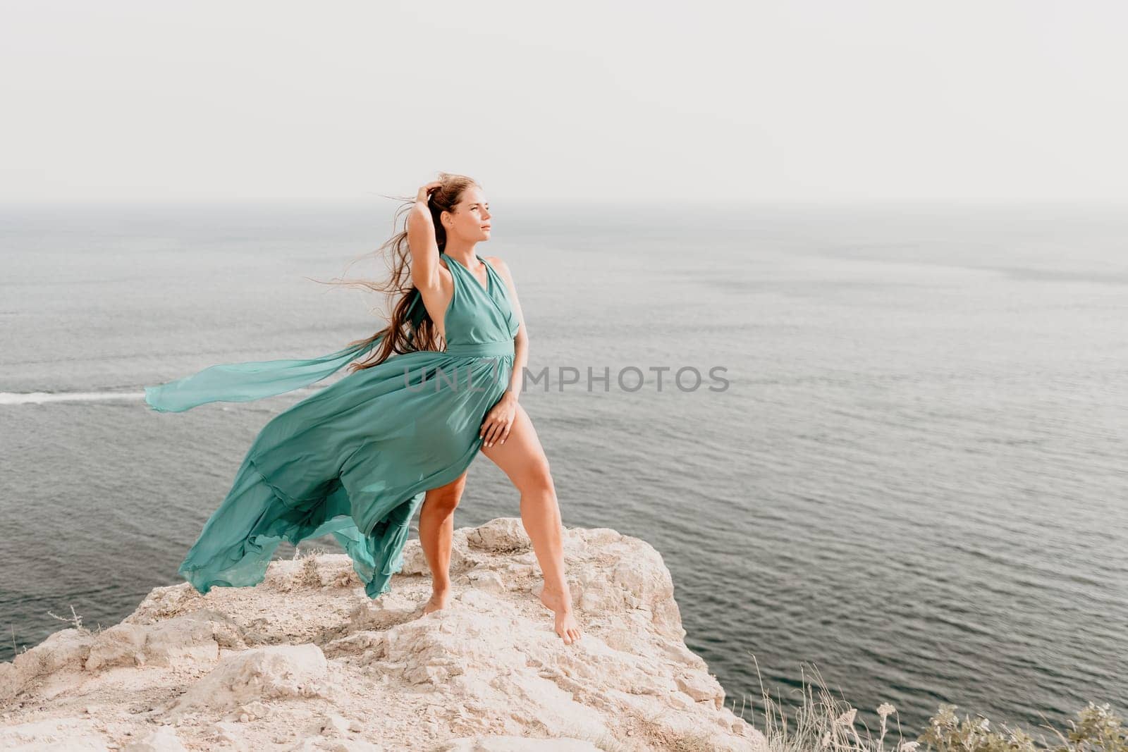 Woman travel portrait. Happy woman with long hair looking at camera and smiling. Close up portrait cute woman in a mint long dress posing on a volcanic rock high above the sea by panophotograph