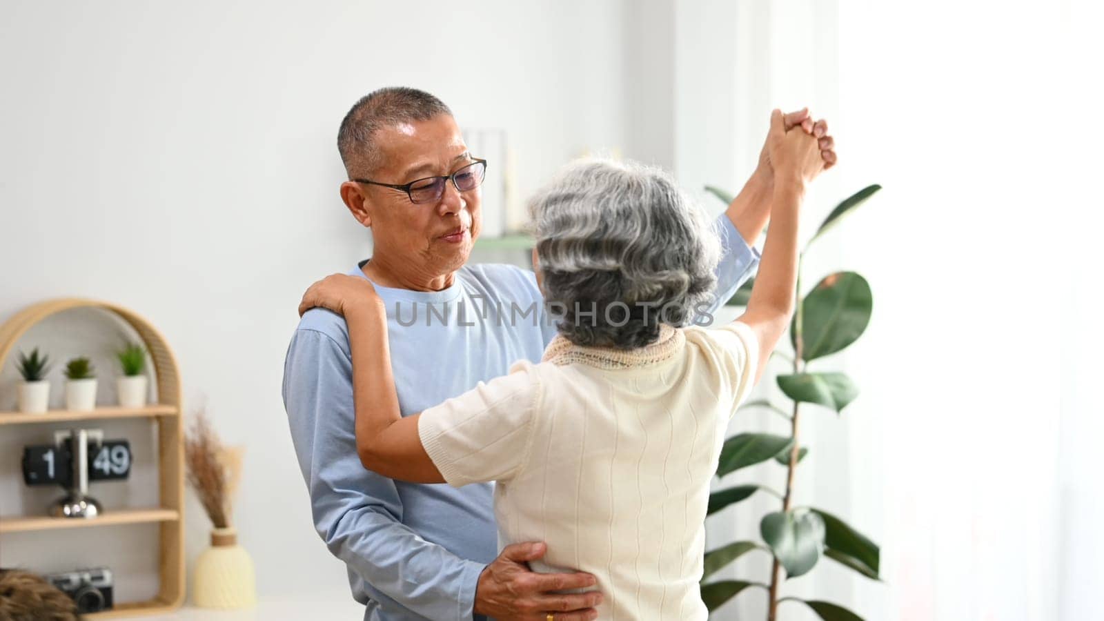 Affectionate senior couple dancing in living room, enjoying retired life and spending free time together at home by prathanchorruangsak