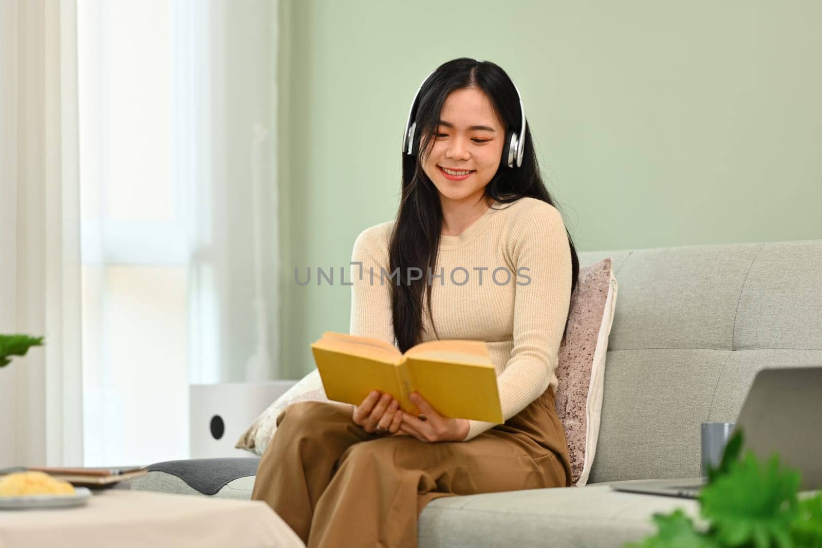 Portrait of smiling young asian woman in headphone reading book on couch. Education, leisure and lifestyle concept.