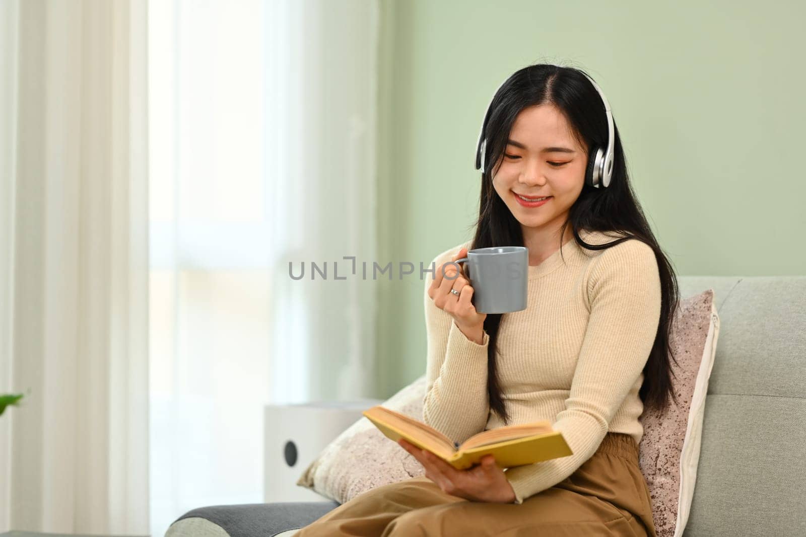 Carefree young woman in headphone drinking coffee and reading book on couch.