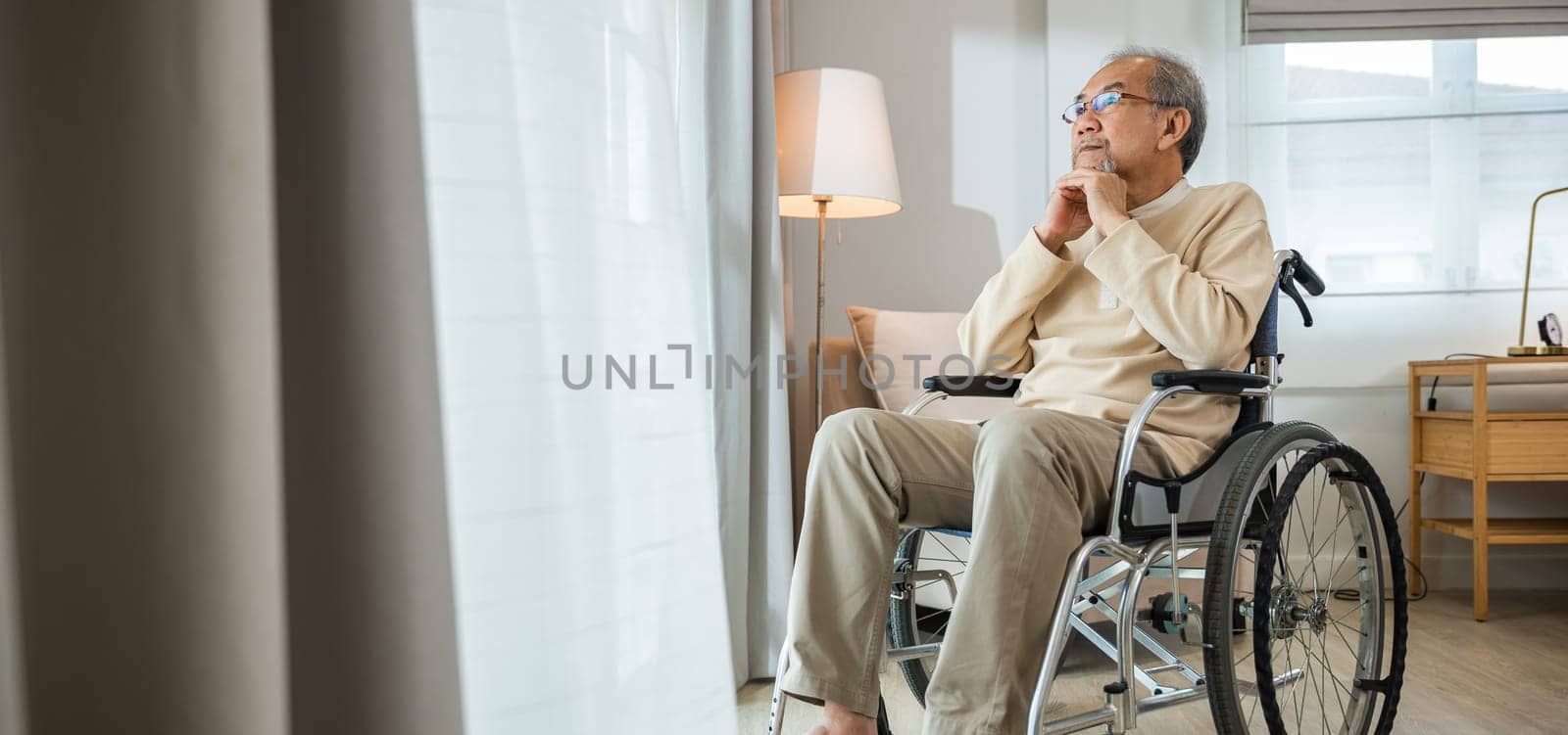 Asian senior man disabled sitting alone in wheelchair looking through window at hospital, lonely elder thoughtful sad old man look outside windows in bedroom at retirement home, feel depressed lonely