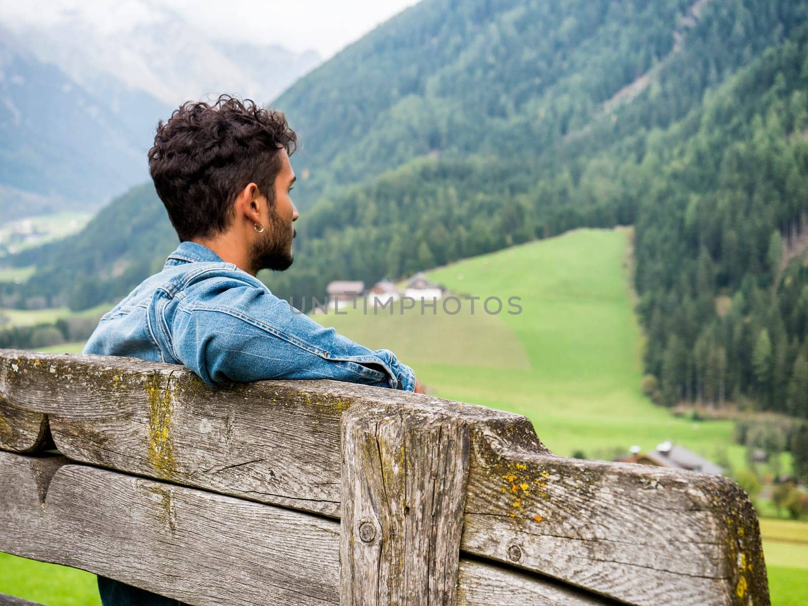 A man sitting on a wooden bench overlooking a valley. Photo of a man enjoying the scenic view from a rustic wooden bench in the valley