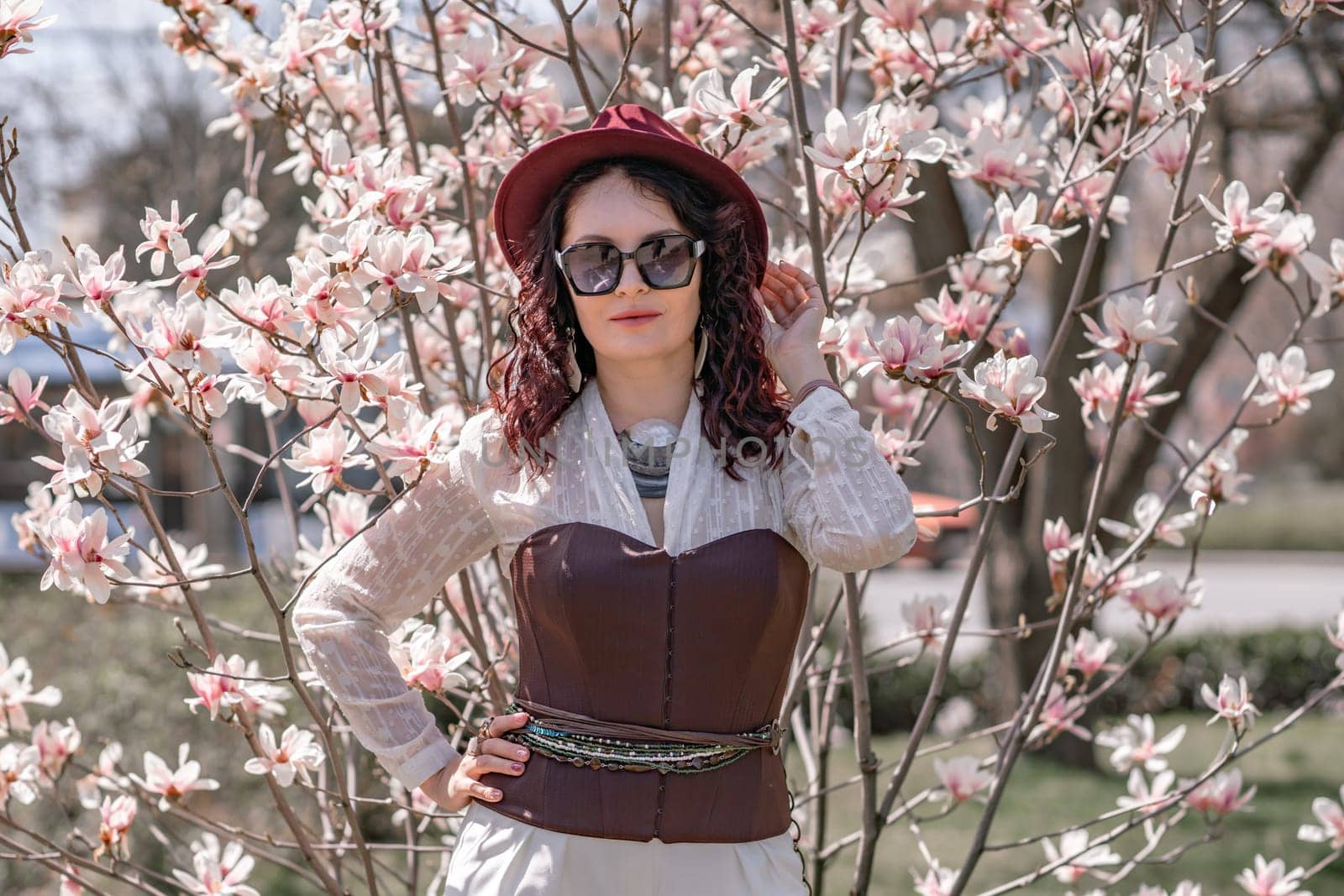 Magnolia park woman. Stylish woman in a hat stands near the magnolia bush in the park. Dressed in white corset pants and posing for the camera