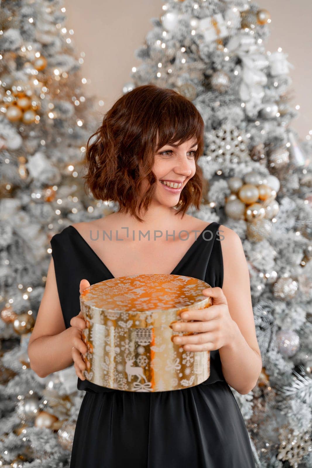 Woman Christmas tree gift. Happy romantic brunette girl in black dress standing on the background of the Christmas tree. Cheerful lady was surprised by the gift after opening the gift box. Merry Christmas and Happy Holidays by Matiunina