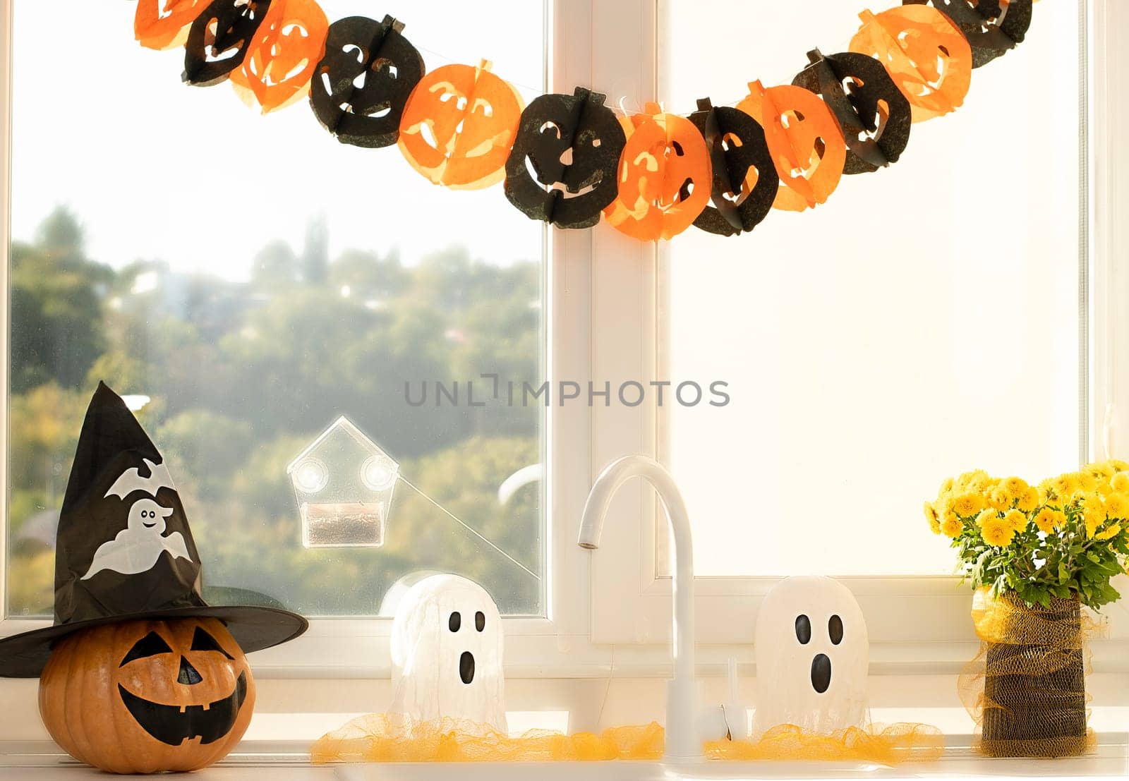 Halloween holiday concept. A pumpkin with a painted face, a white ghost and a bouquet of yellow chrysanthemum flowers in a black vase against the background of a window in home interior.