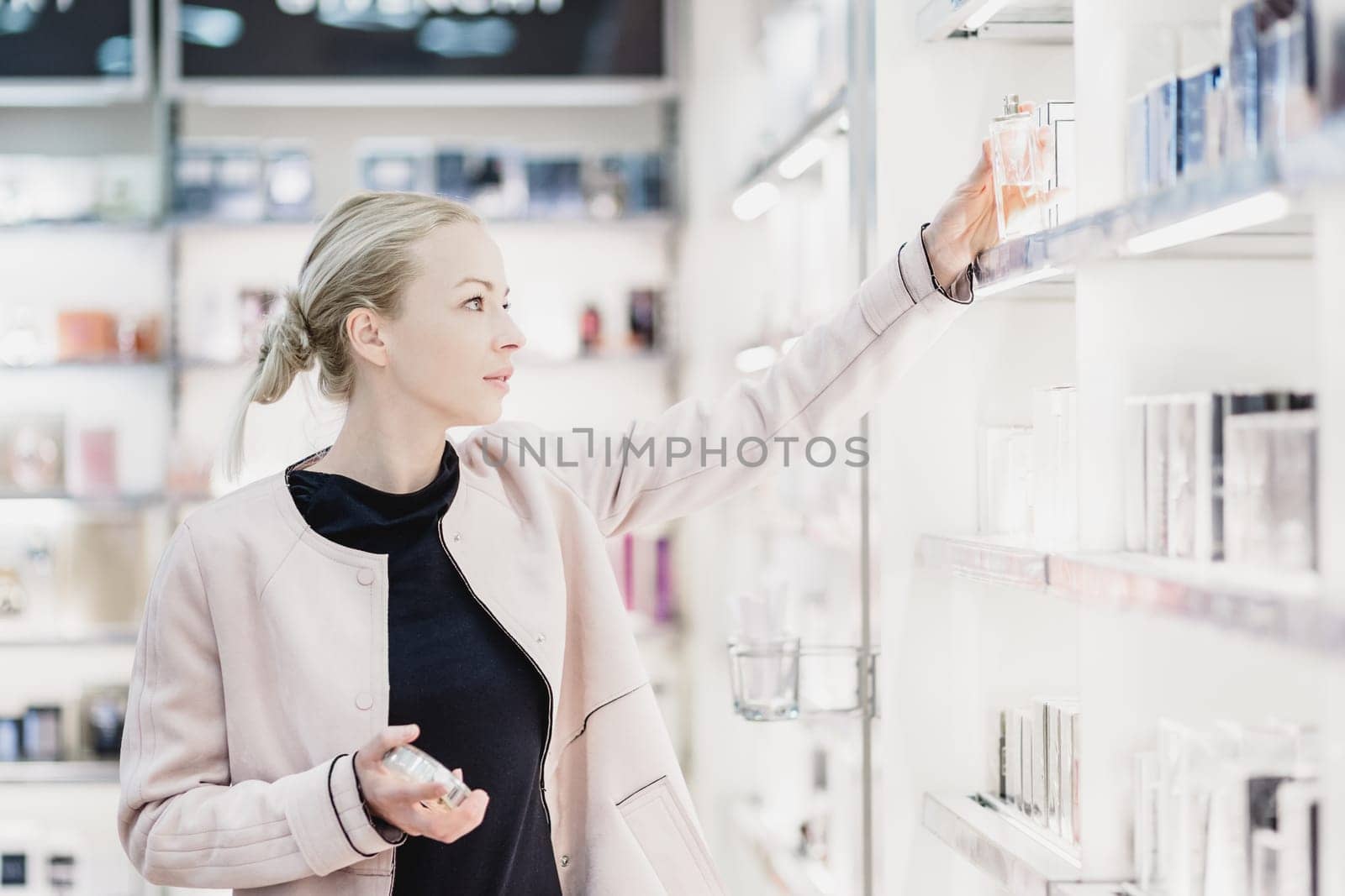 Elegant blond young woman choosing perfume in retail store. Beautiful blond lady testing and buying cosmetics in a beauty store.