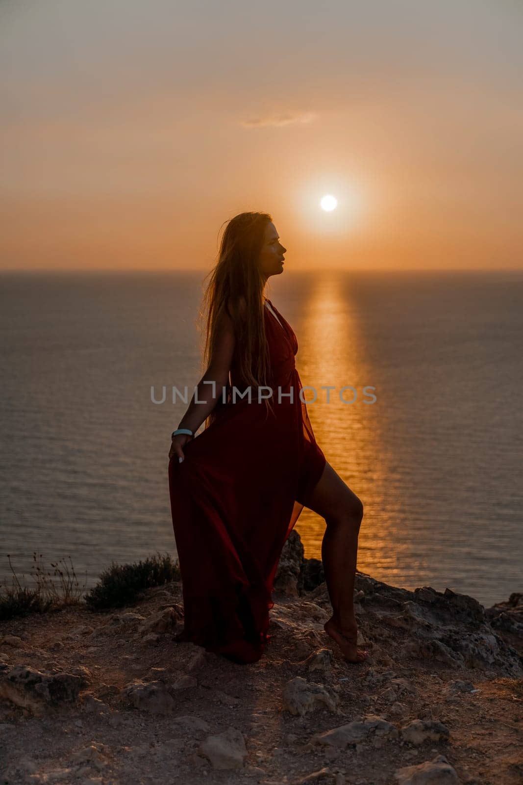 Woman sunset sea red dress, back view a happy beautiful sensual woman in a red long dress posing on a rock high above the sea on sunset