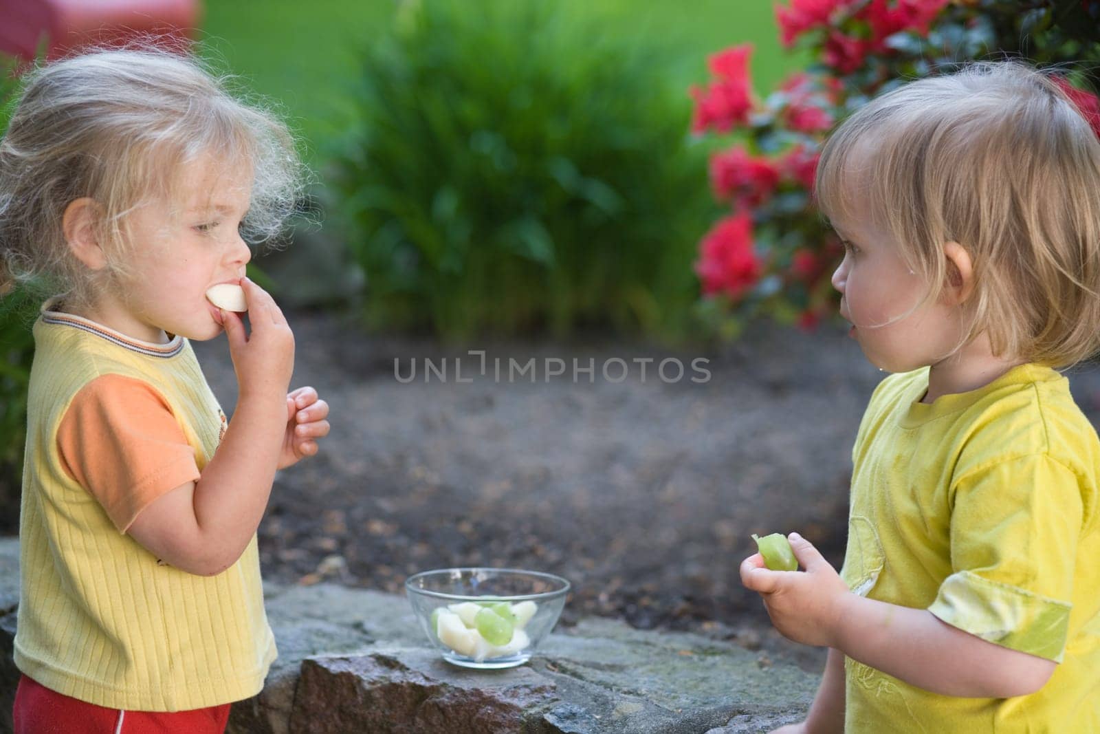 Two little girls stand outside against a wall and eat fruit with relish