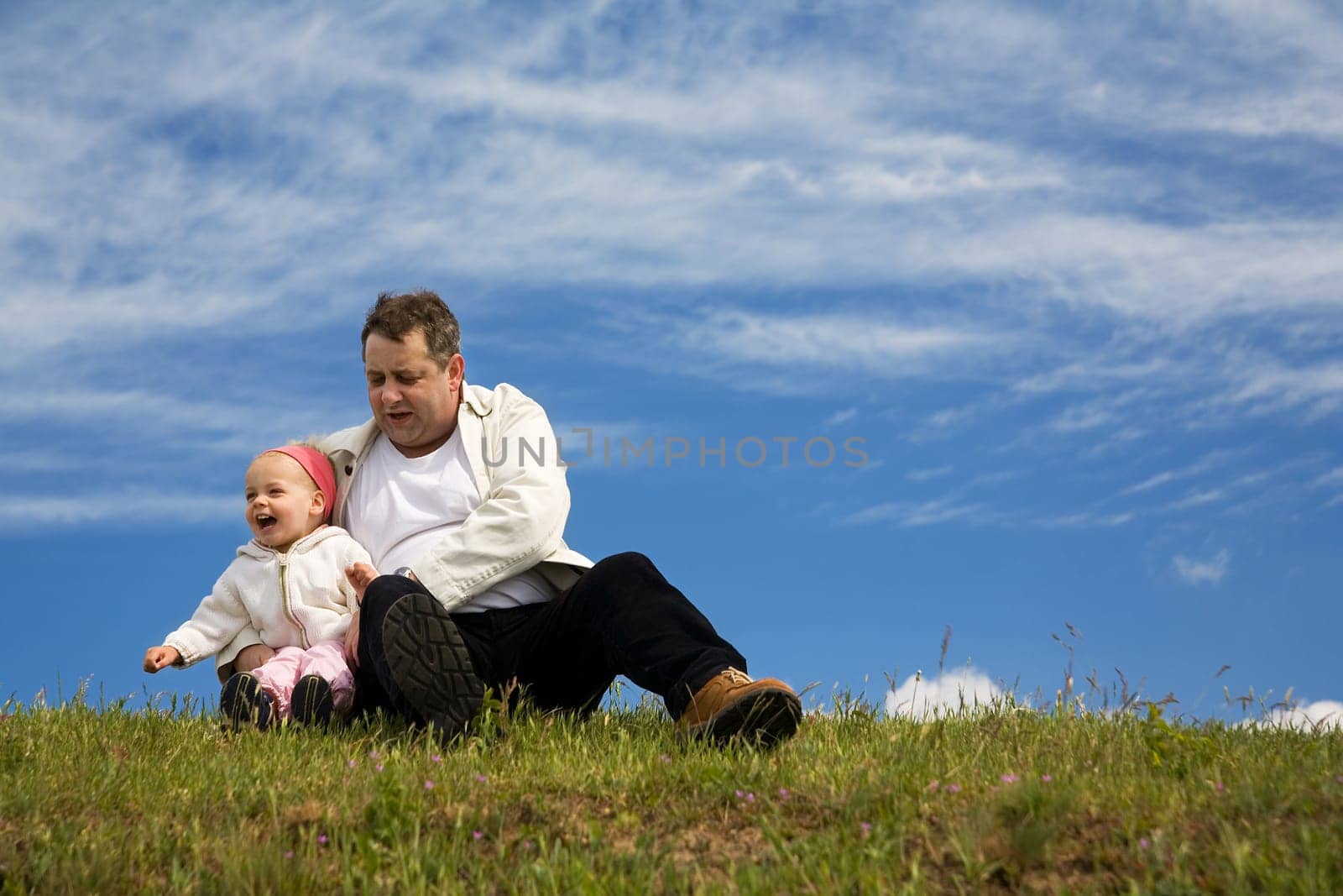 Father and daughter sitting in a meadow in front of a blue,slightly cloudy sky