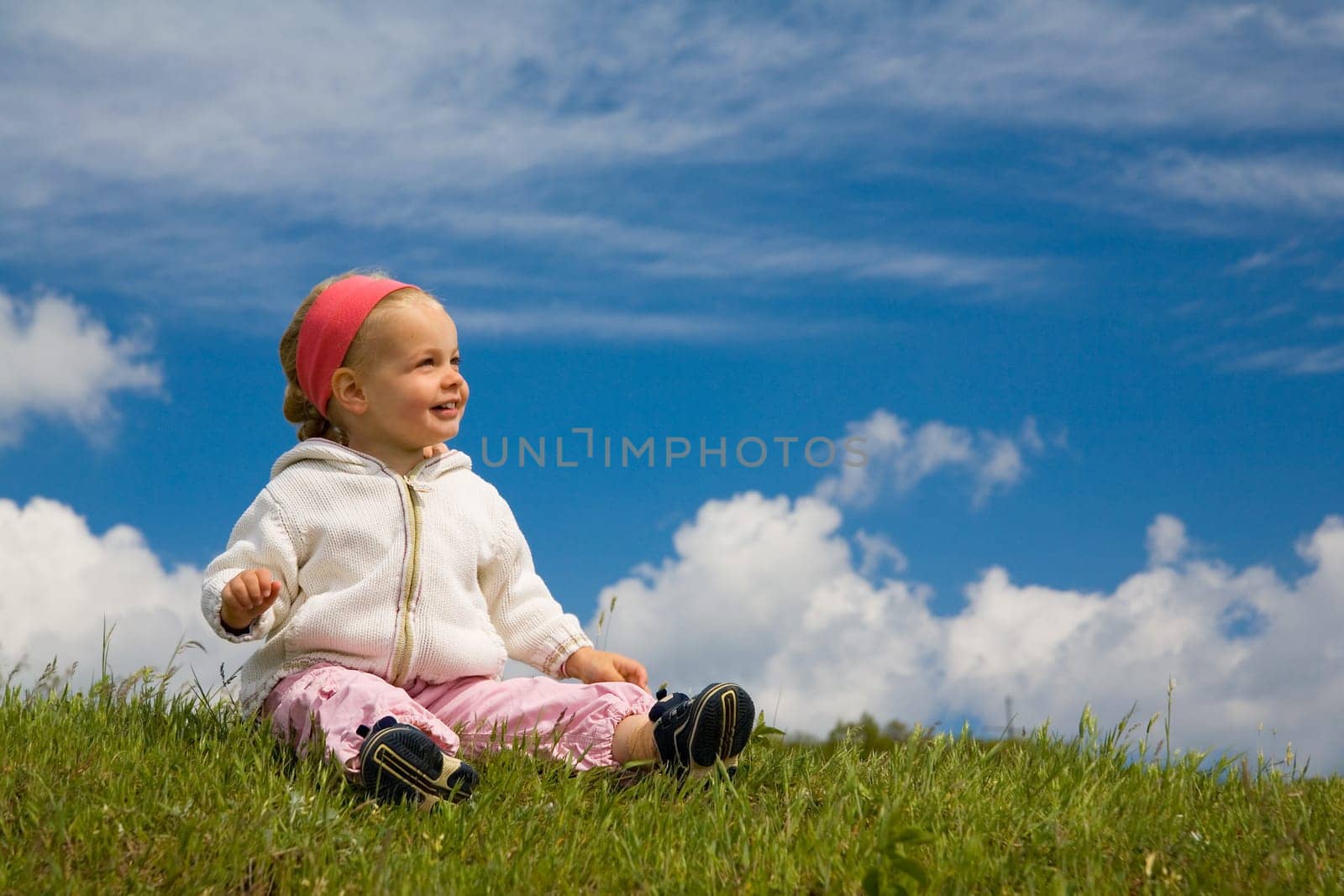 A little girl sits on a summer meadow against a blue slightly cloudy background