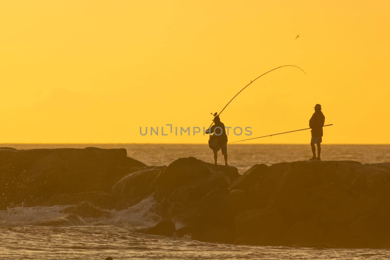 Fishermen are fishing standing on the rocks in the sea at sunset by Studia72