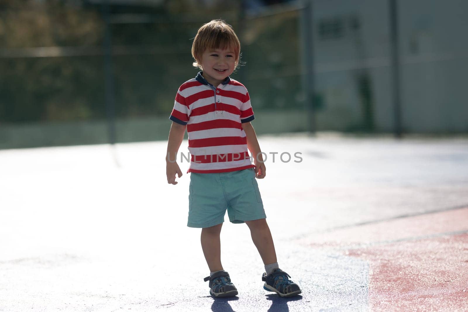 Little blonde smiling boy standing on the outdoors sports playground. Mid shot