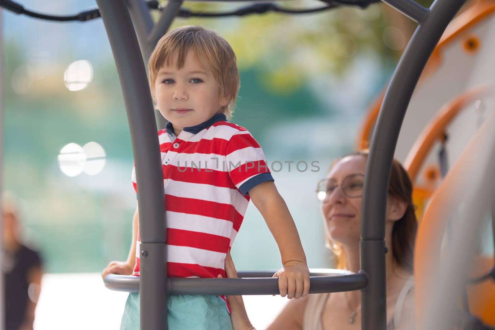 Cute blond boy climbing ladder on playground with support of his smiling mother - the boy looks in the camera by Studia72