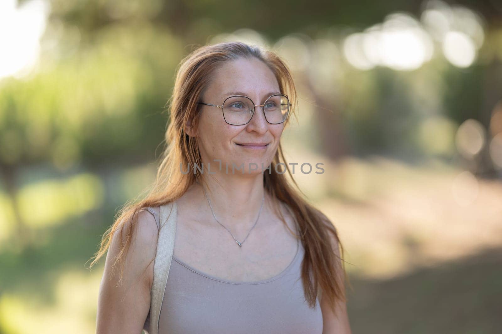 Charming adult woman in glasses with dyed red hair standing in the park looking forwards. Portrait