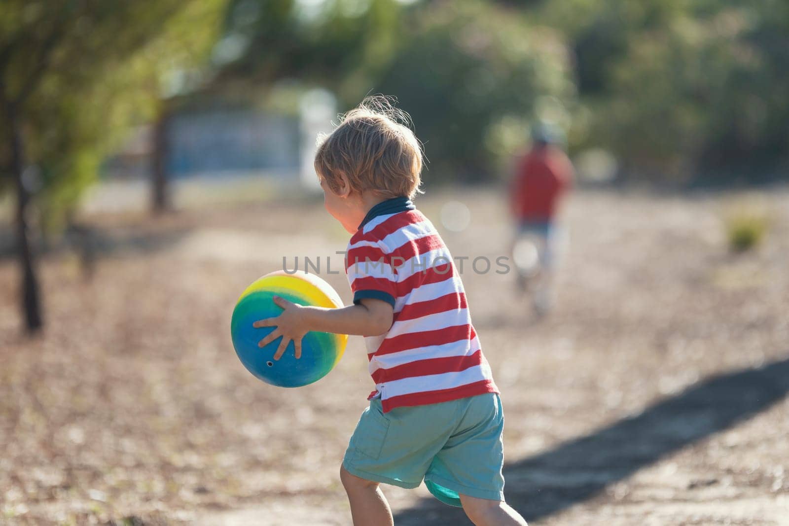Little blonde boy running forwards in the park holding a ball. Mid shot