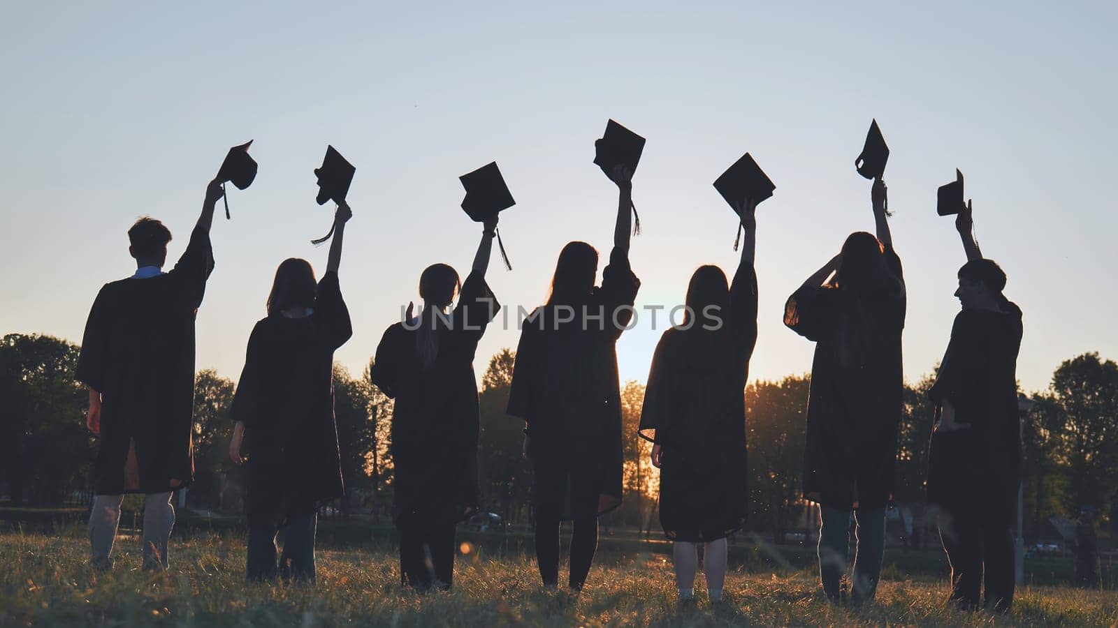 Silhouettes of college graduates waving their caps at sunset. by DovidPro