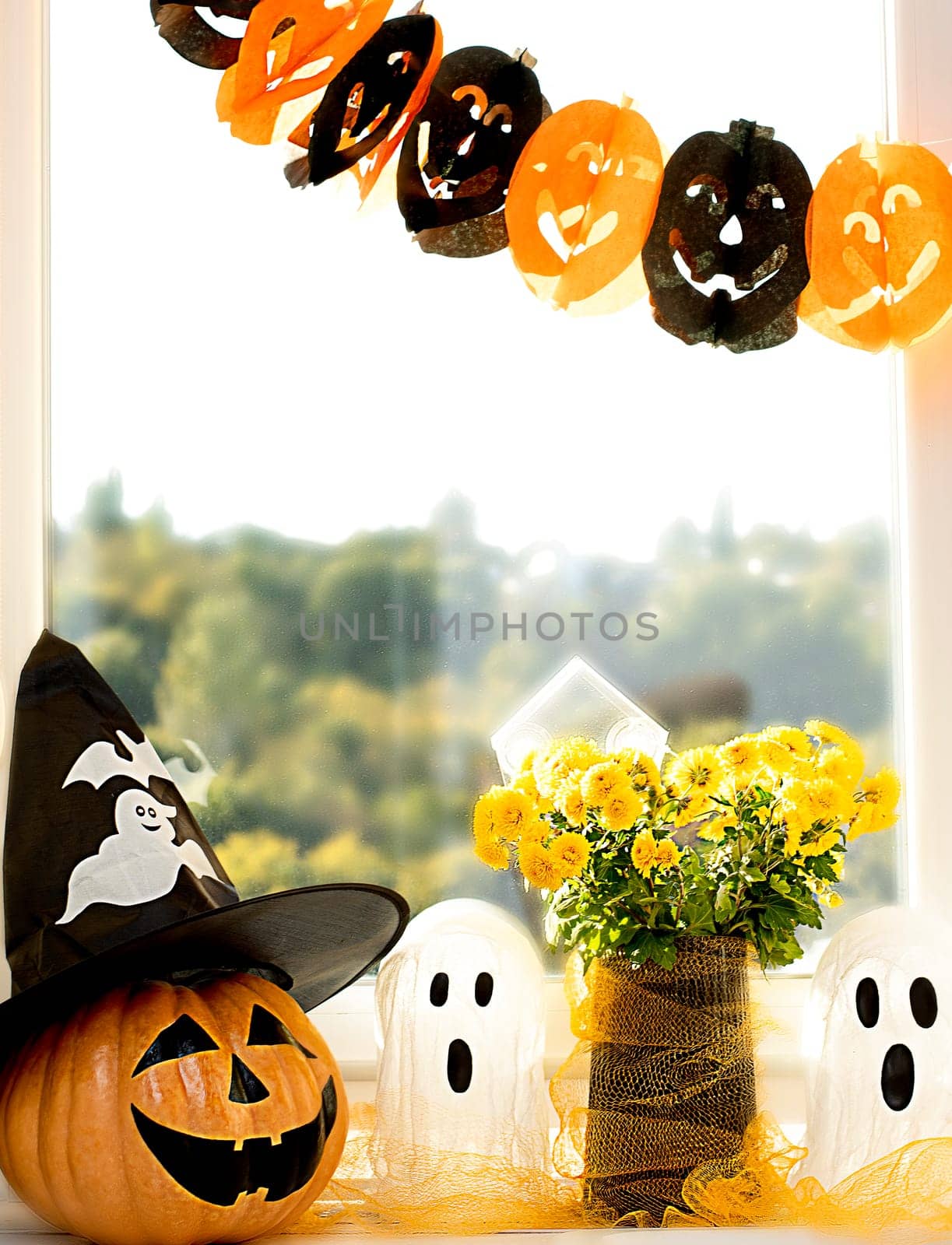 Halloween concept. A pumpkin with a painted face, a white ghost and a bouquet of yellow chrysanthemum flowers in a black vase against the background of a window in home interior. Close-up, vertical.