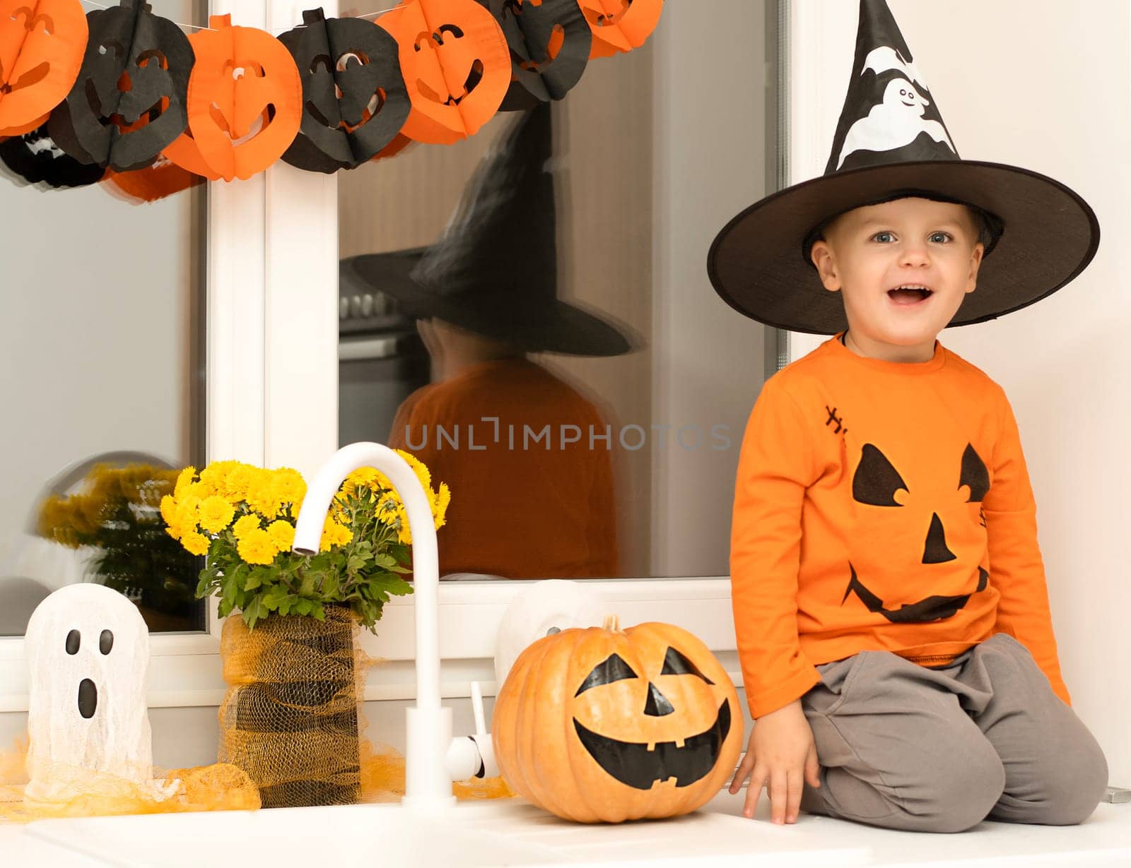 Halloween concept. A small, cheerful, handsome boy in a wizard's hat and an orange sweater sits on a table in the kitchen against the background of a pumpkin, a ghost and a bouquet of yellow flowers. Festive interior.