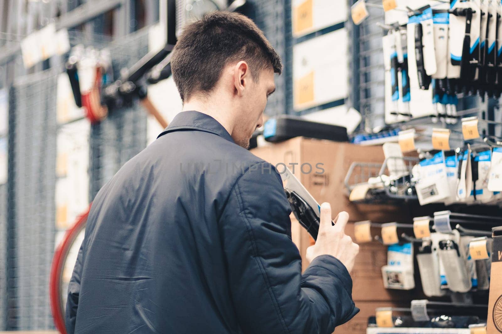 One young caucasian guy in a blue jacket stands sideways at the blurry counters in the store and examines the goods, choosing a purchase, close-up side view.