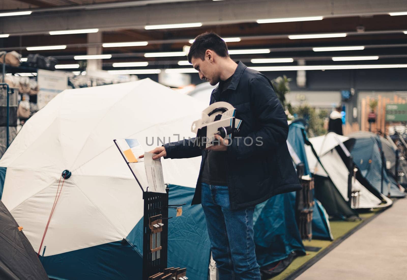 One young Caucasian man in a blue jacket stands sideways and reads the instructions for a tent in a store, close-up side view.