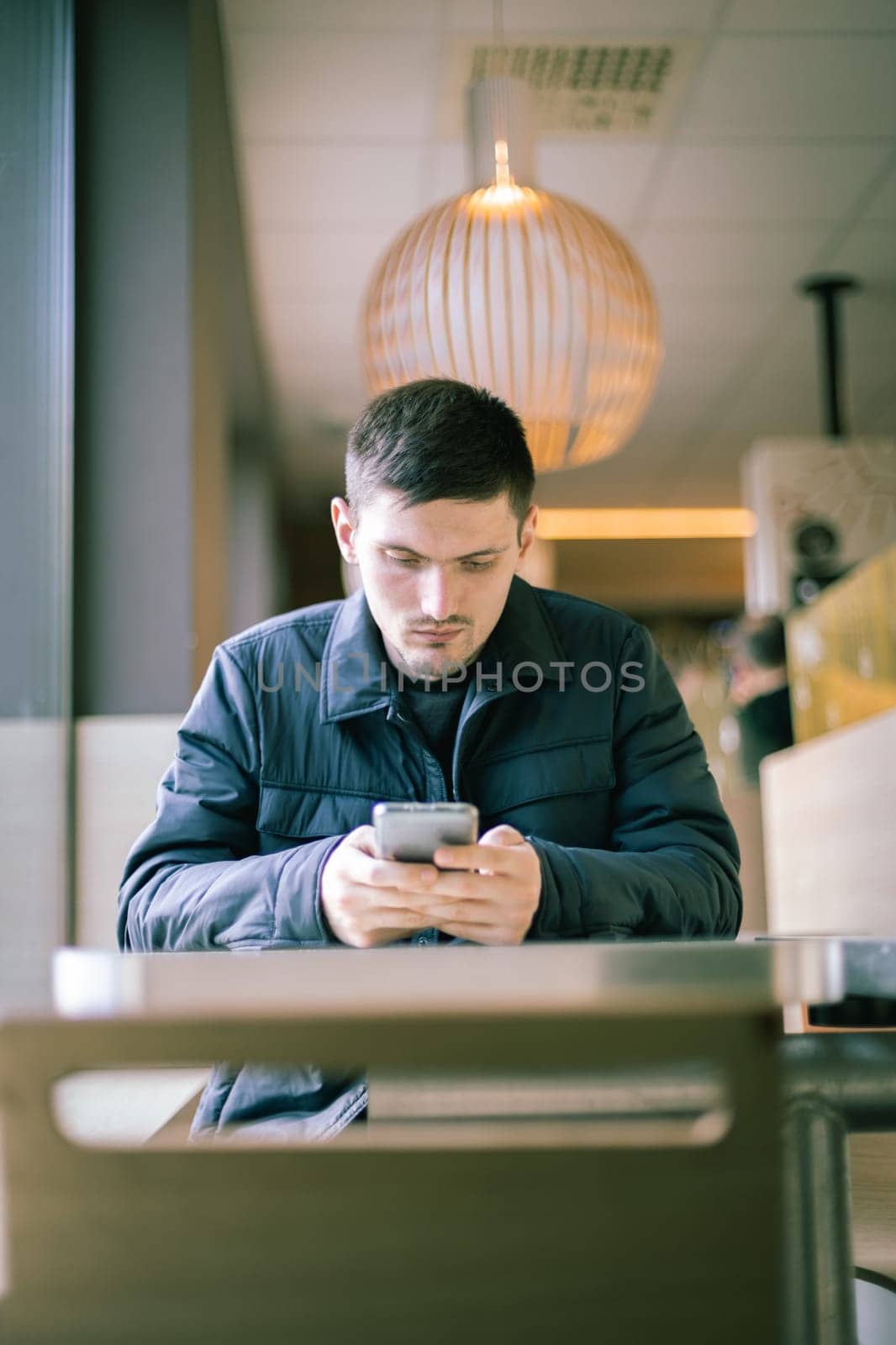 One young Caucasian man in a dark jacket sits at a table by the window in a diner and carefully reads a message in a mobile phone in his hands, close-up view from below.