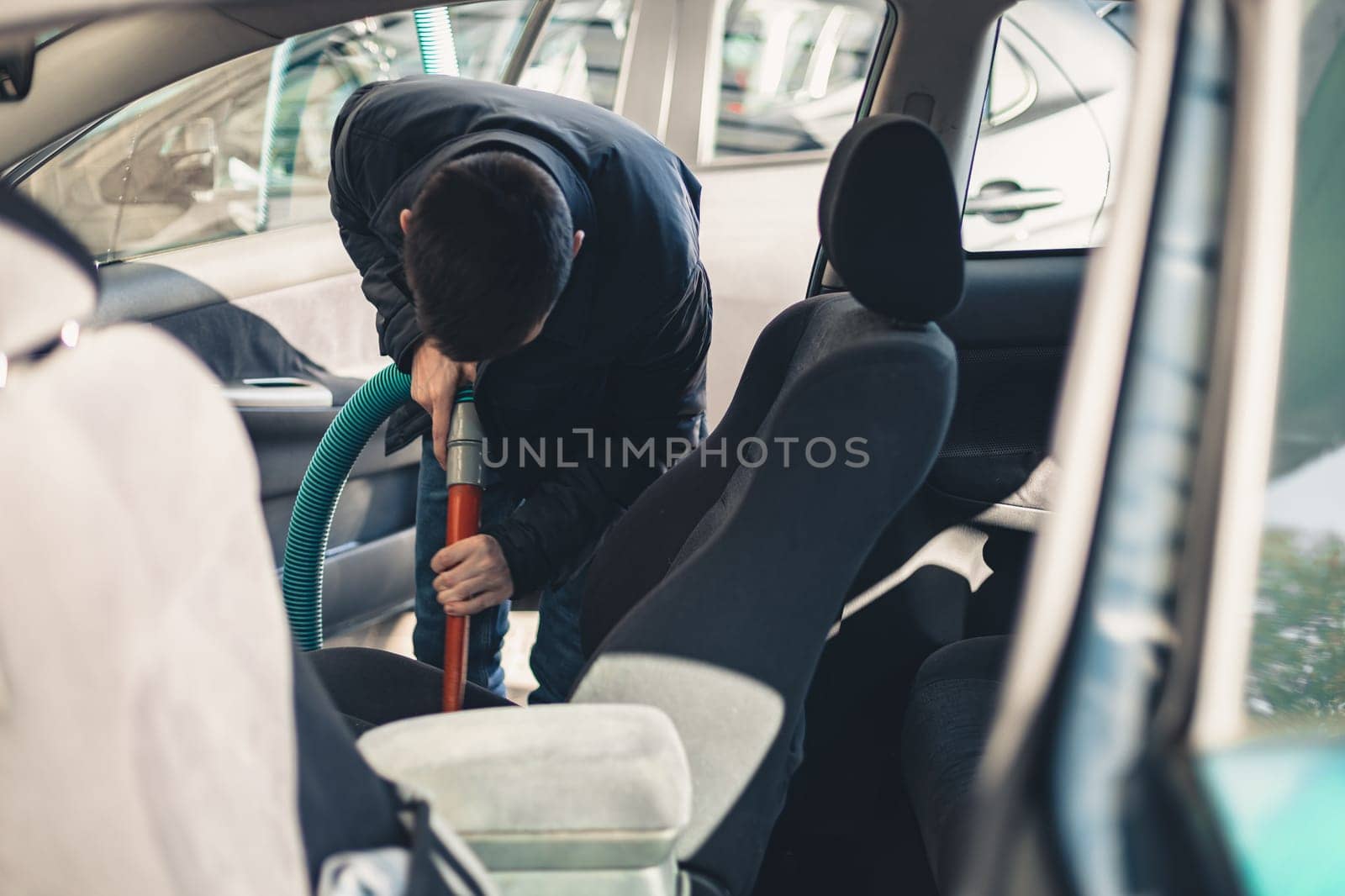 One young handsome caucasian brunette man vacuuming the front passenger seat inside the salon in his car at the city car wash on a clear spring sunny day, top view, close-up.