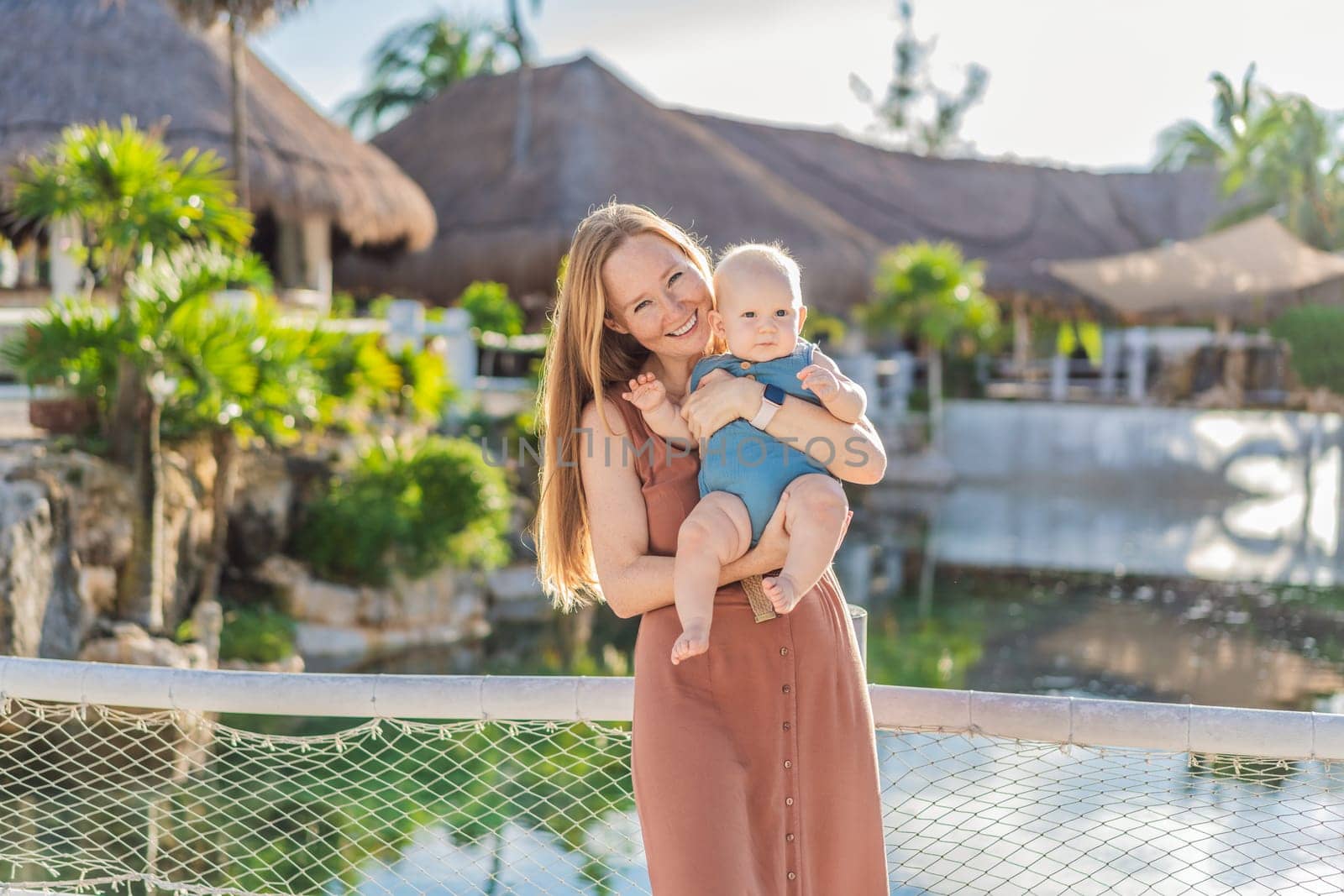 Amidst tropical palms and thatched roofs, a loving mom embraces her baby, sharing warmth and affection in a tranquil outdoor setting by galitskaya