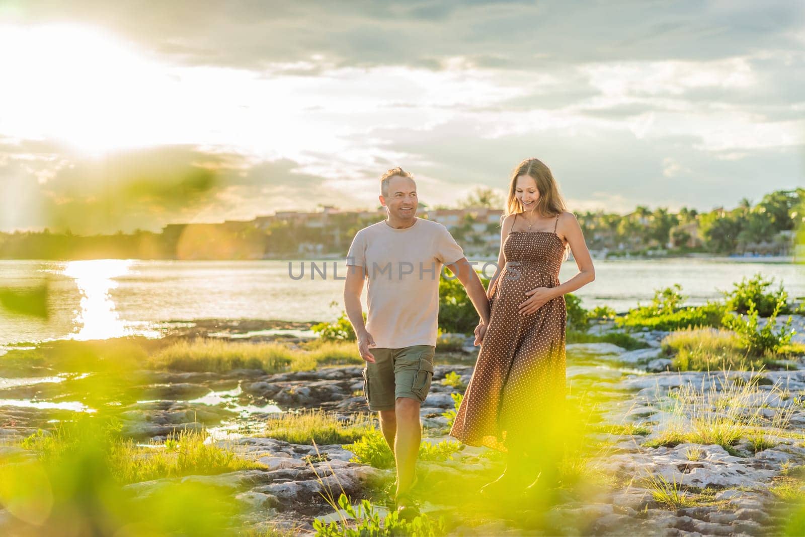 A happy, mature couple over 40, enjoying a leisurely walk on the waterfront On the Sunset, their joy evident as they embrace the journey of pregnancy later in life.