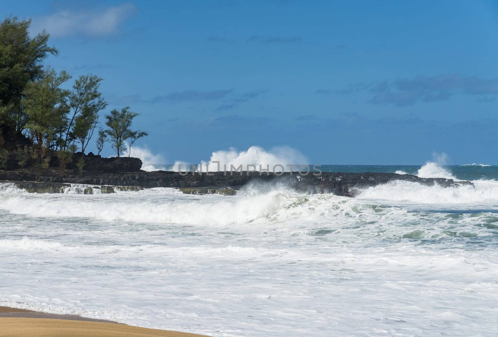 Ocean waves crashing on the shore rocks under a blue sky, with the turquoise water rippling and splashing at Lumahai Beach.