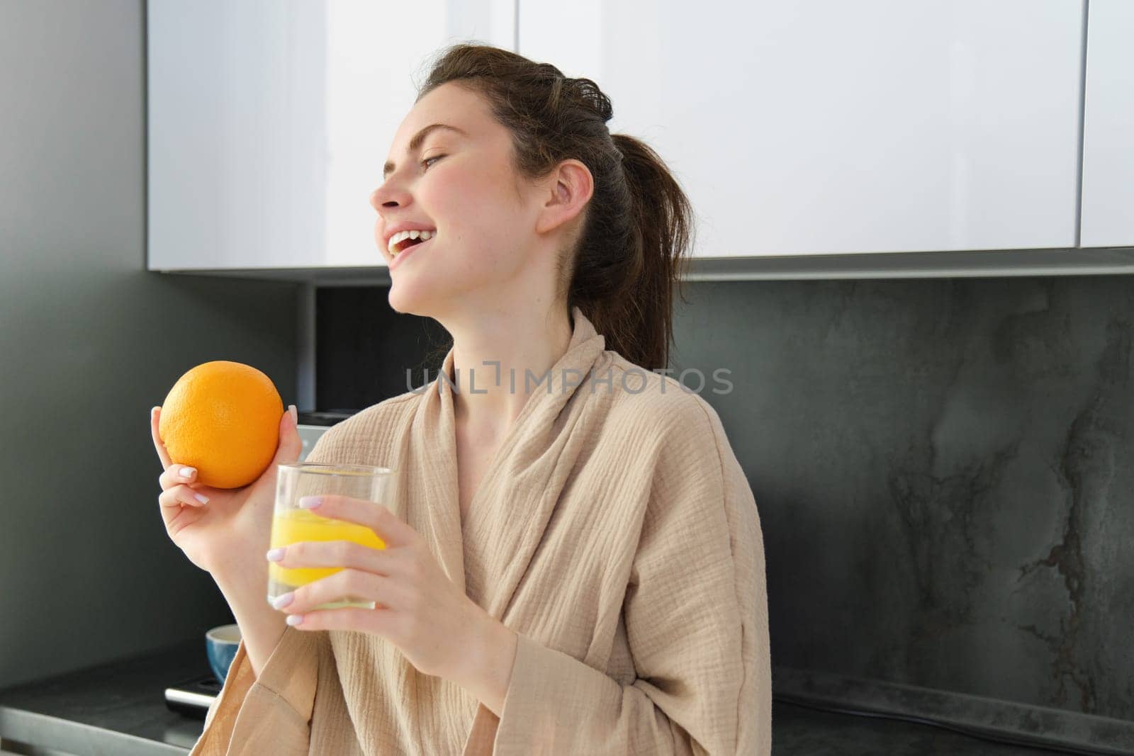 Portrait of happy woman in kitchen, wearing bathrobe, drinking orange juice, freshly squeezed drink, smiling and laughing, food and drink concept.