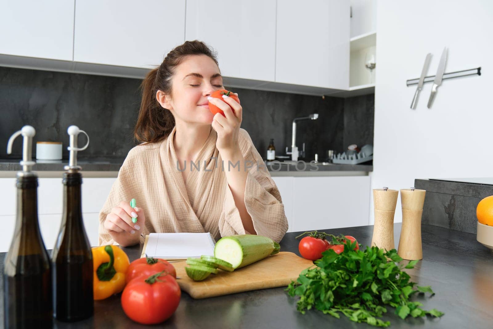 Portrait of beautiful young woman in the kitchen, writing down cooking recipe, sitting near chopping board with vegetables and making grocery list, creating healthy veggie menu for her family.