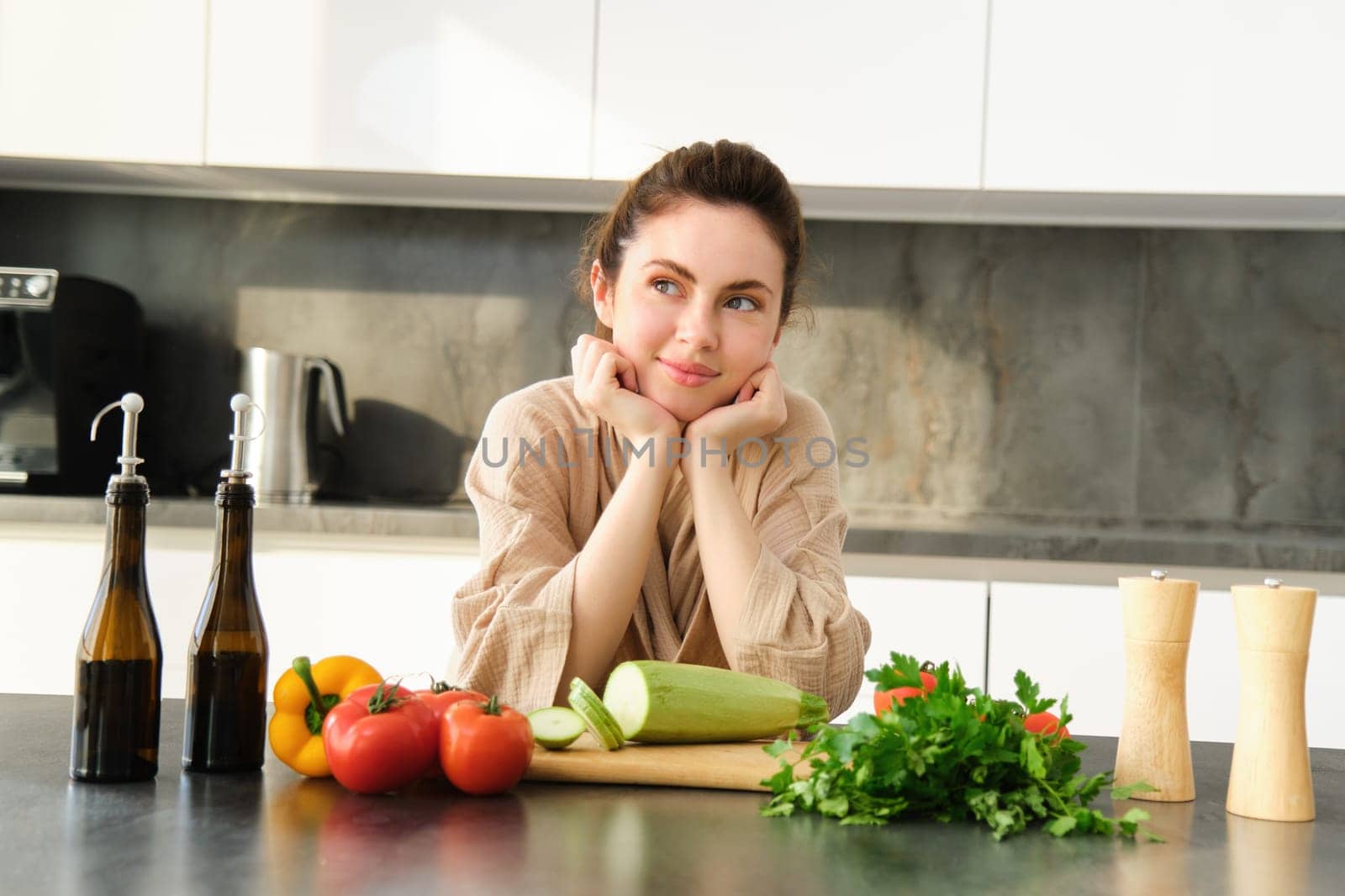 Portrait of smiling young woman leaning on kitchen counter, preparing dinner, standing near chopping board, cutting zucchini, looking away and thinking, thinks what to cook.