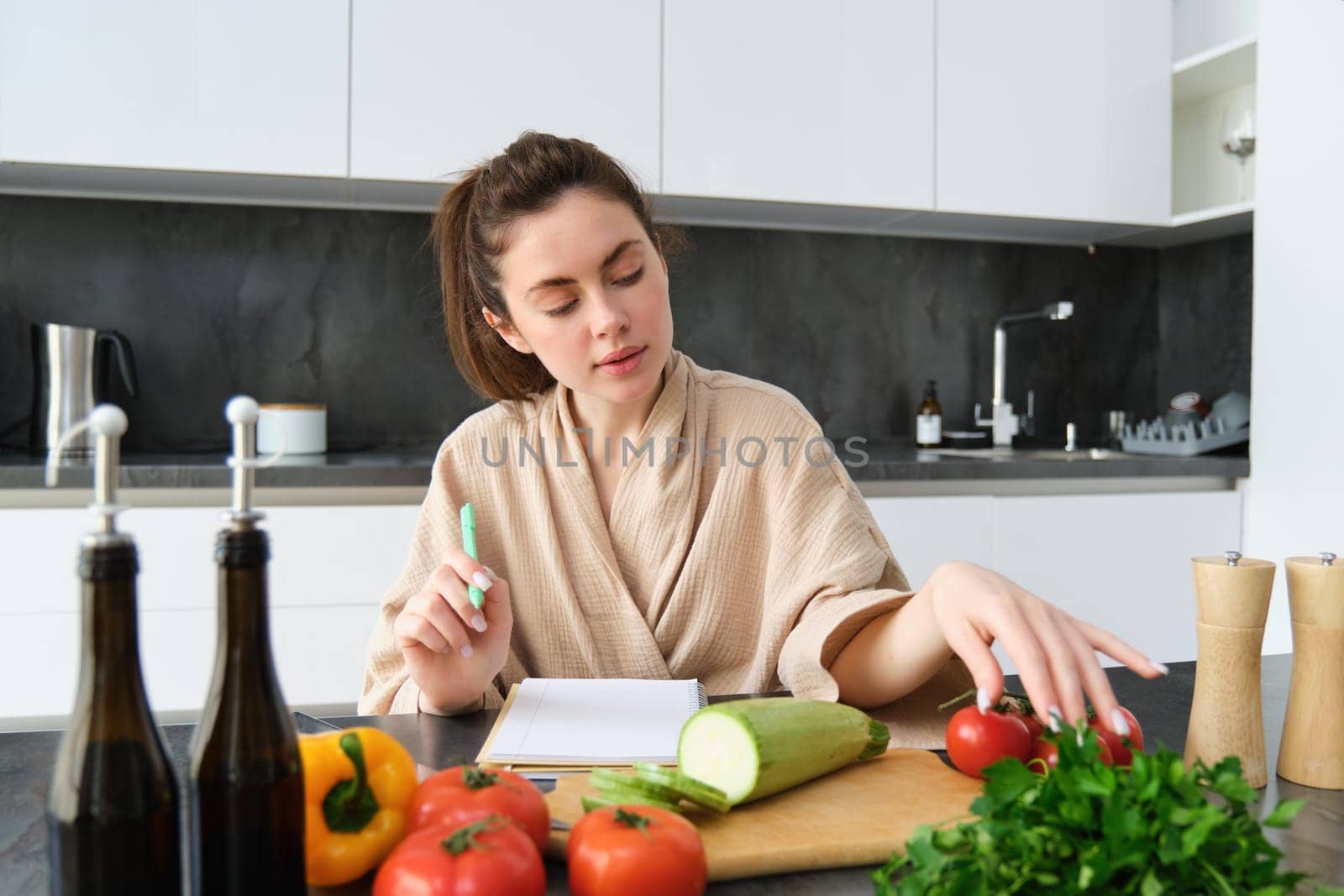 Portrait of woman cooking in the kitchen, sitting in front of vegetables, tomatoes zucchini and parsley, making list of groceries, writing down recipe, wearing bathrobe.
